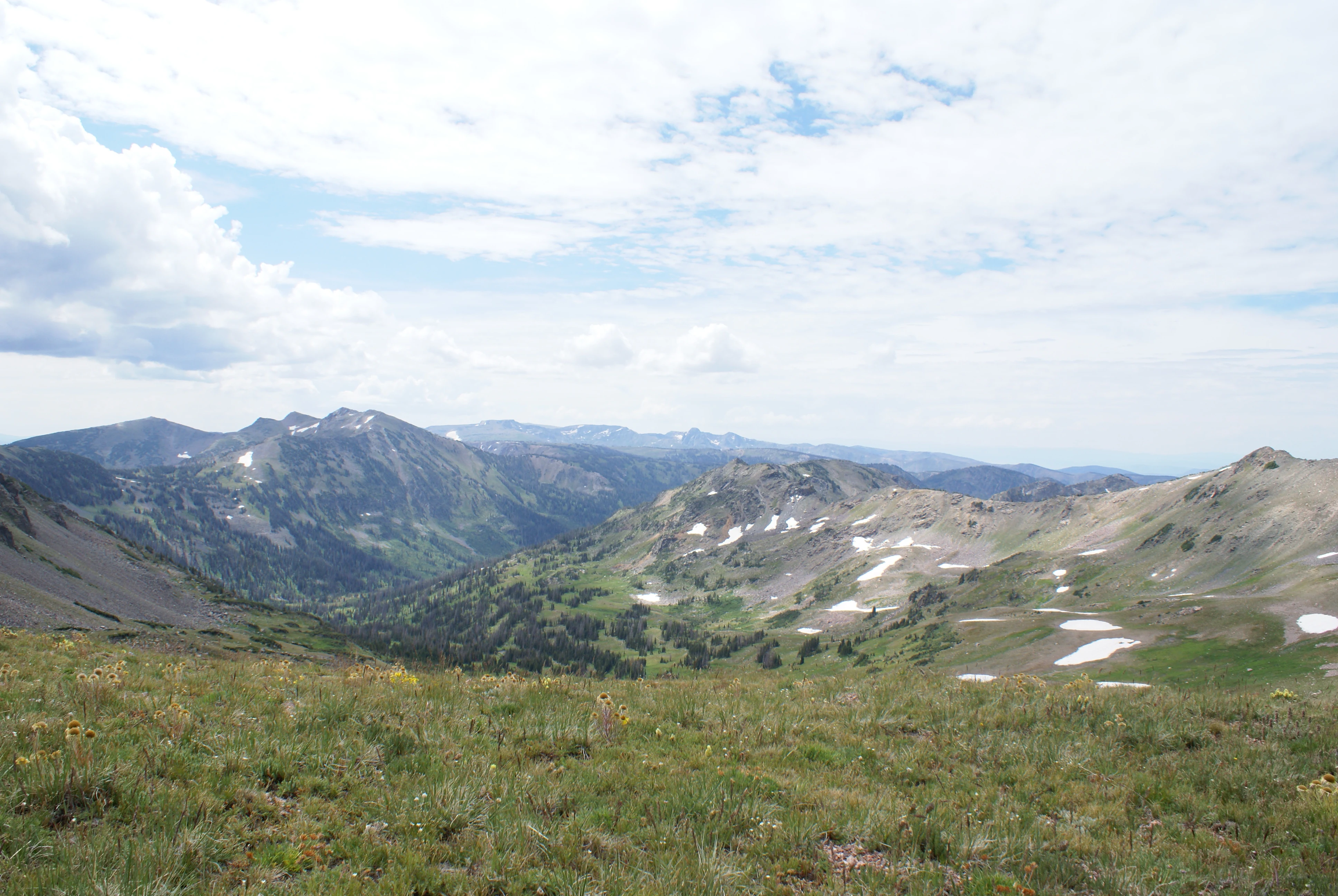 A wide-angle mountain landscape view featuring grassy slopes in the foreground, scattered patches of snow on the peaks, and a deep valley in the middle distance under a partly cloudy sky.