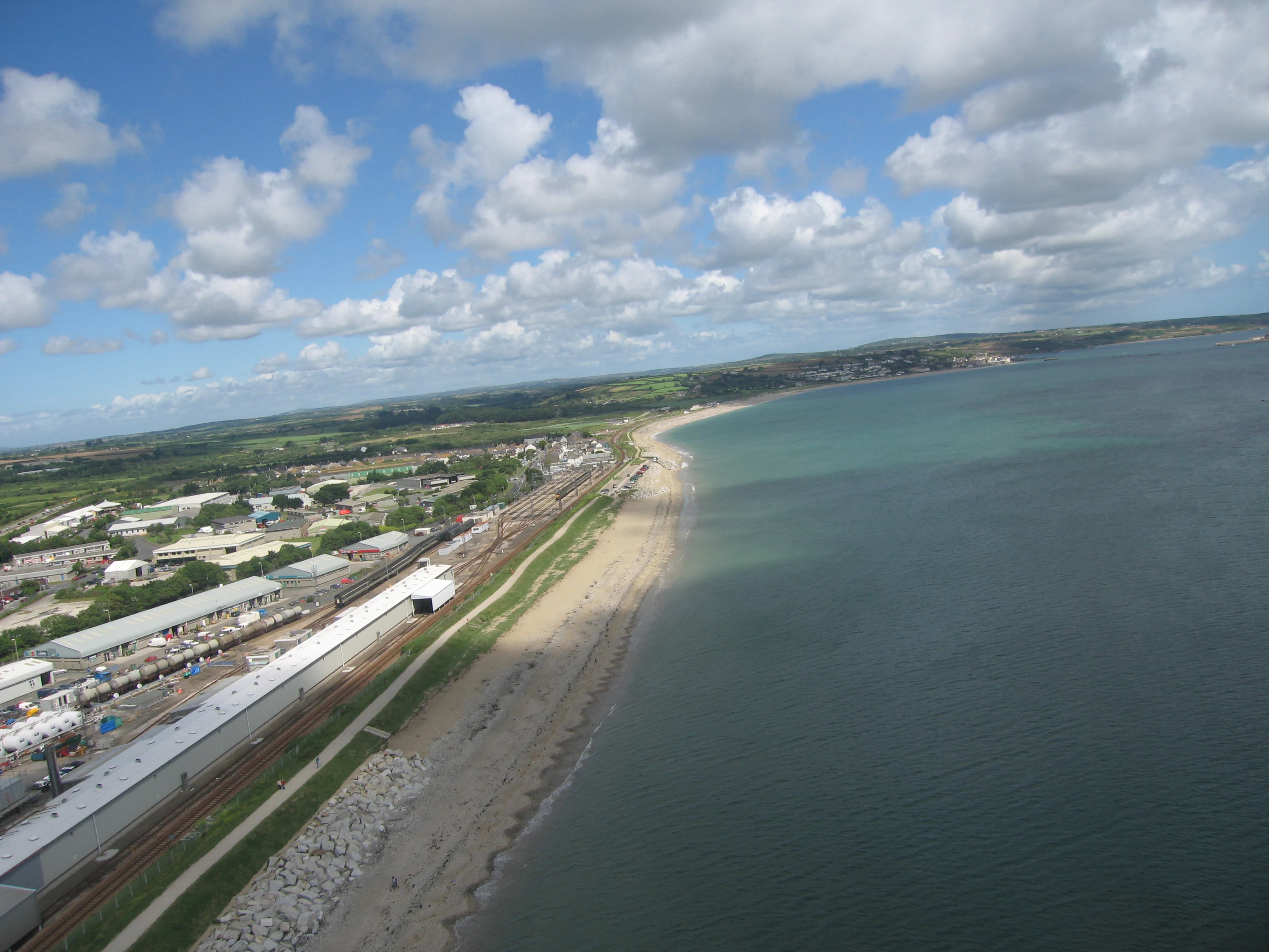 Aerial view of a coastal stretch with a sandy beach meeting the ocean, a railway track running parallel to the shore, and inland development.