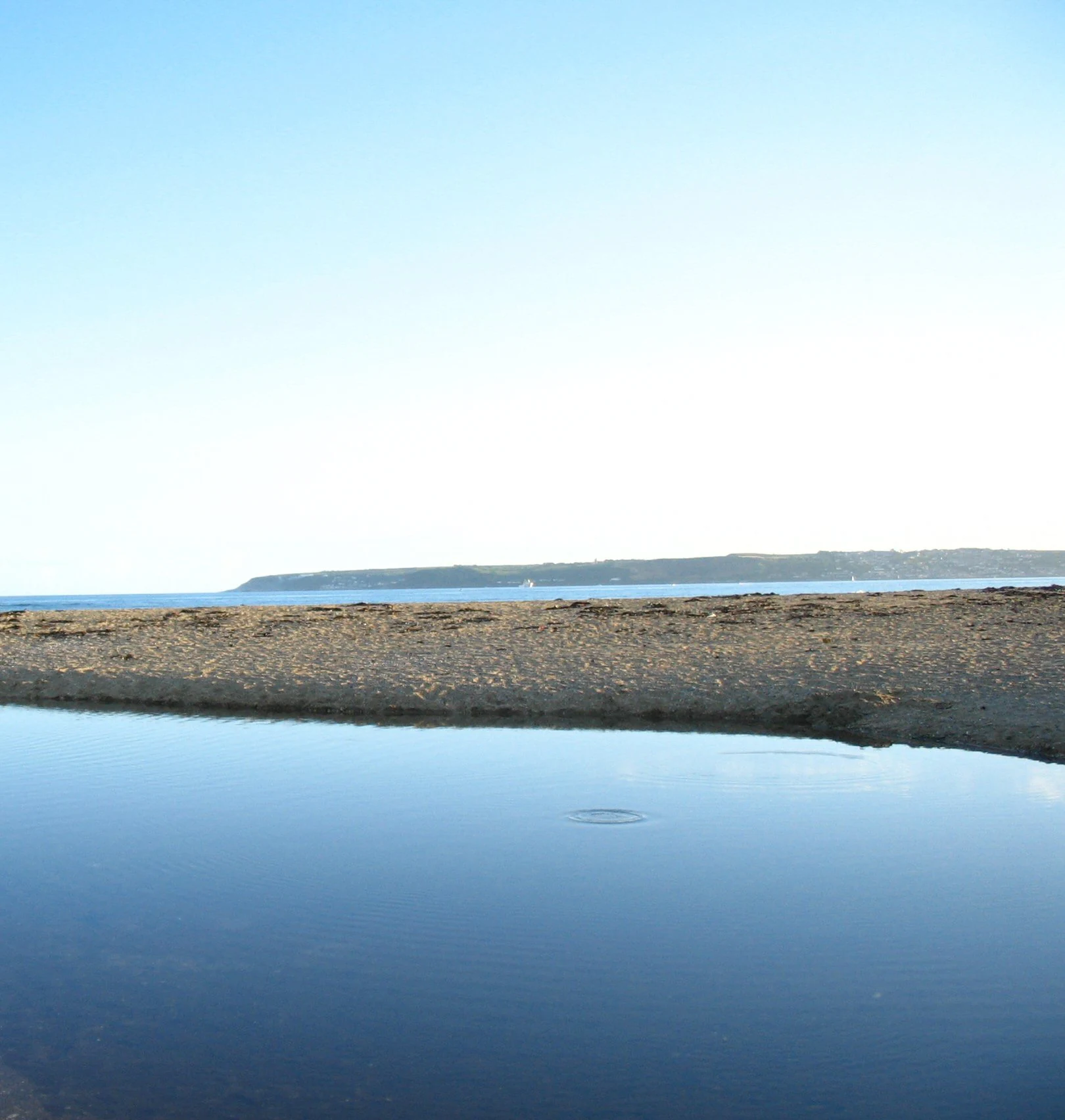 A wide coastal landscape showing a still tidal pool in the foreground with a sandy beach and a distant shoreline across the sea under a clear sky.