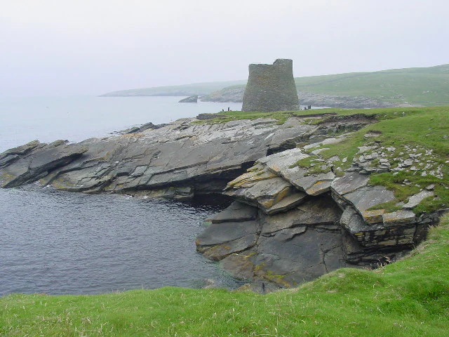 A stone Iron Age tower known as a broch sits on a grassy, rocky coastline overlooking the sea under a misty, overcast sky.