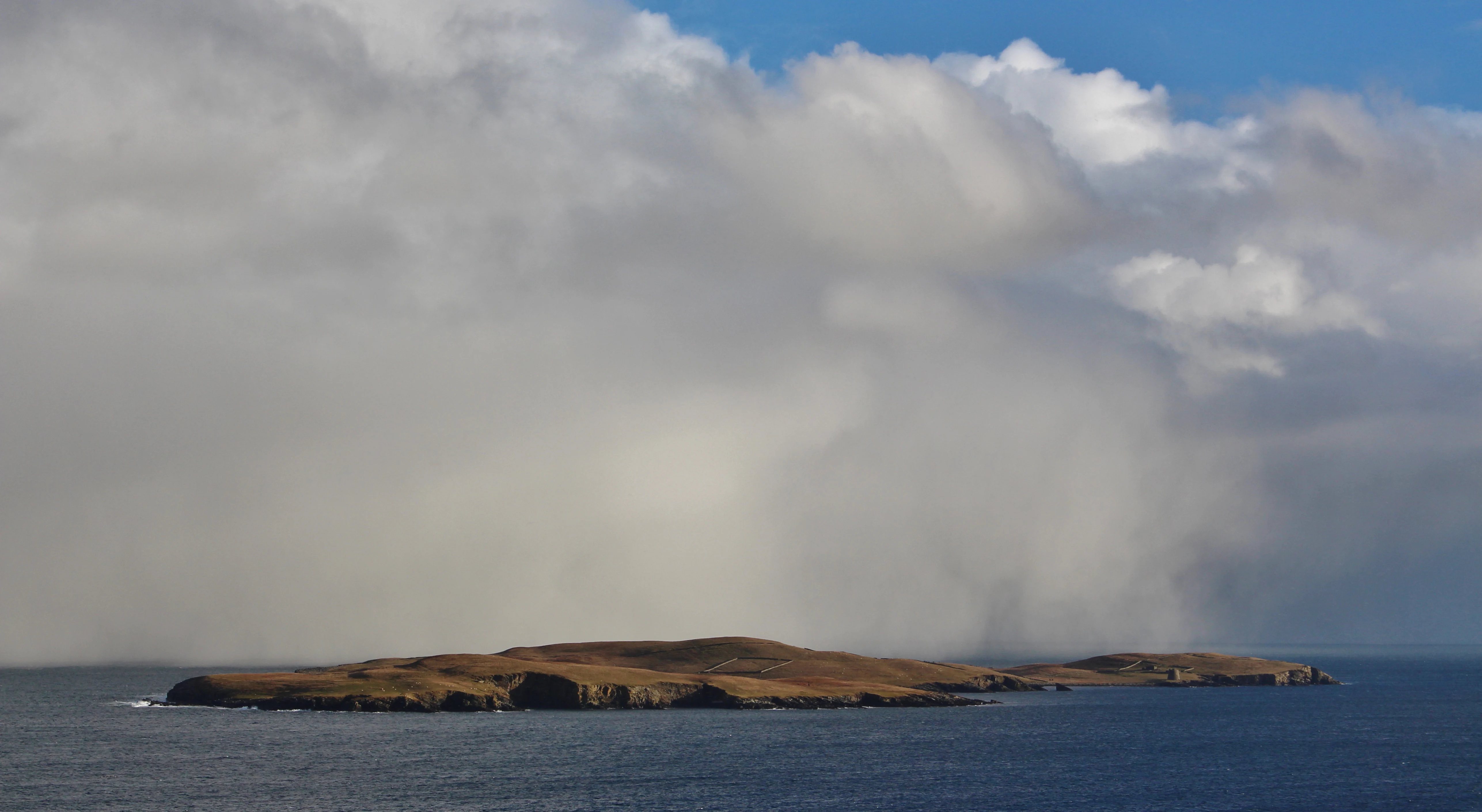 A wide view of the small island of Mousa in the North Sea, partially obscured by heavy white snow or rain clouds under a blue sky.