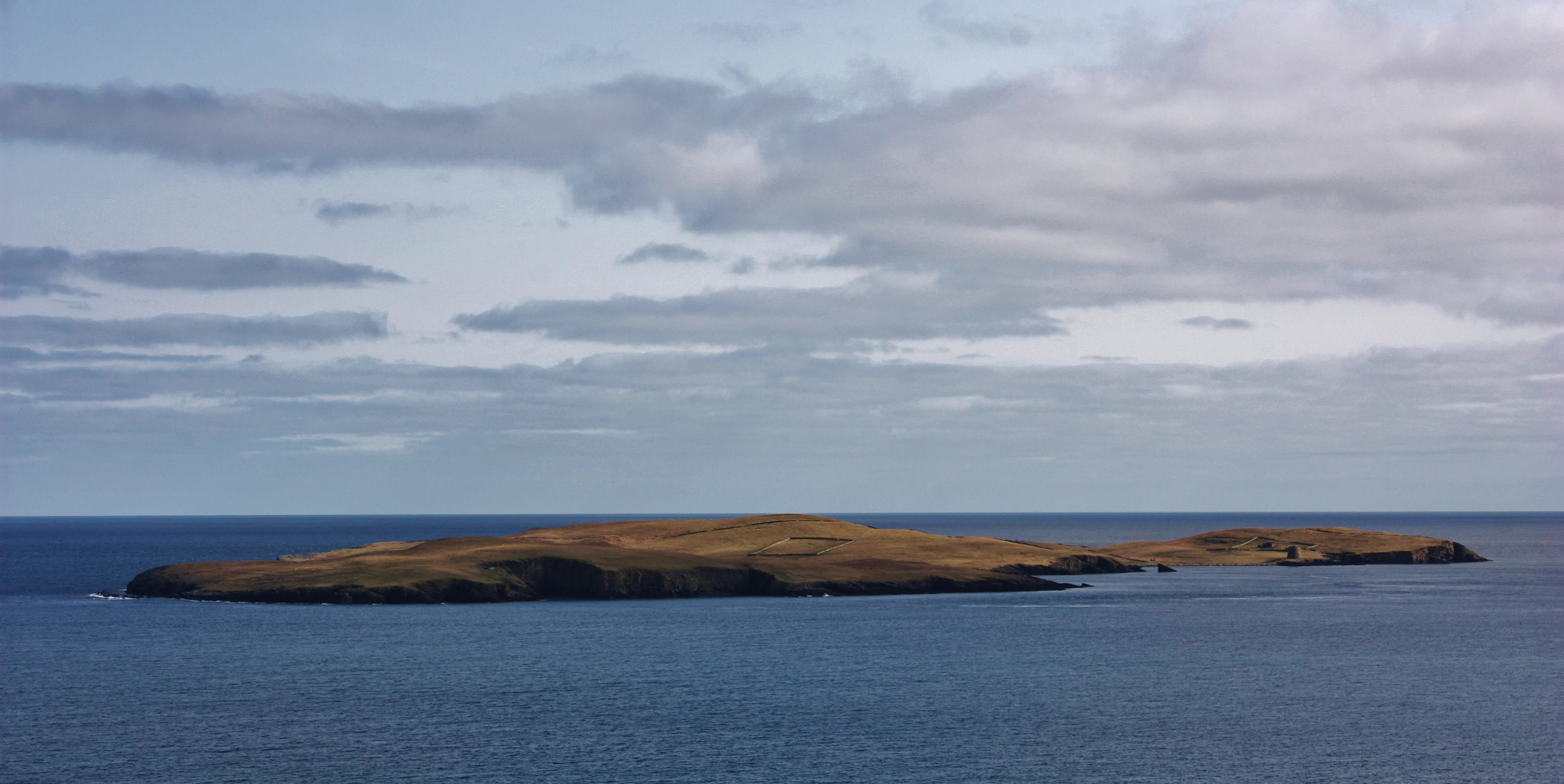 A distant, elevated view of the grassy island of Mousa surrounded by open ocean under a cloudy sky.