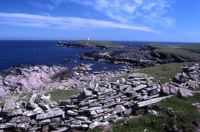 A rocky coastal headland on Mousa island with a stone wall in the foreground and the sea in the background under a blue sky.