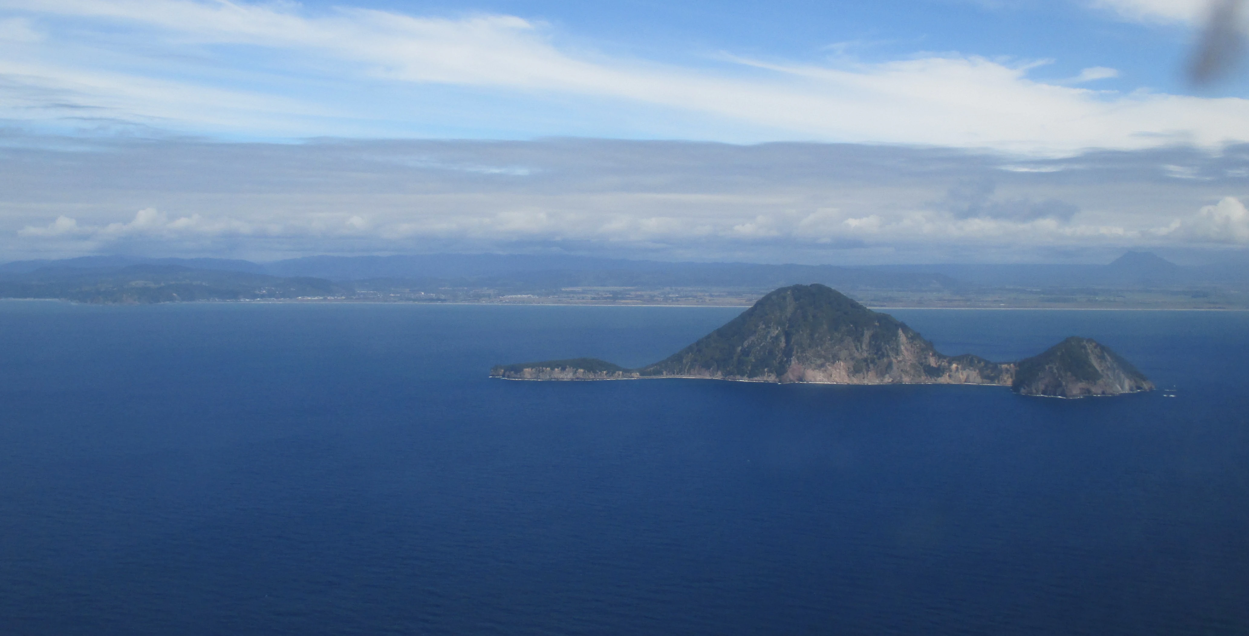 An aerial wide-angle view of the volcanic Moutohora Island surrounded by the open ocean, with a distant coastline visible on the horizon under a cloudy sky.
