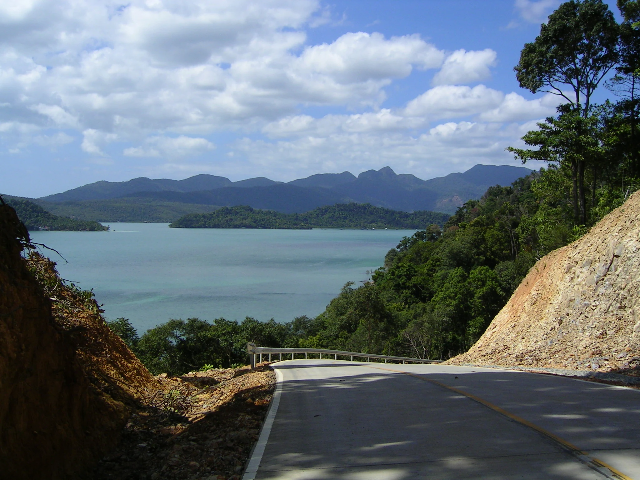 Carretera asfaltada serpentea por la ladera con vistas a una masa de agua con montañas al fondo, rodeada de vegetación exuberante y terreno rocoso