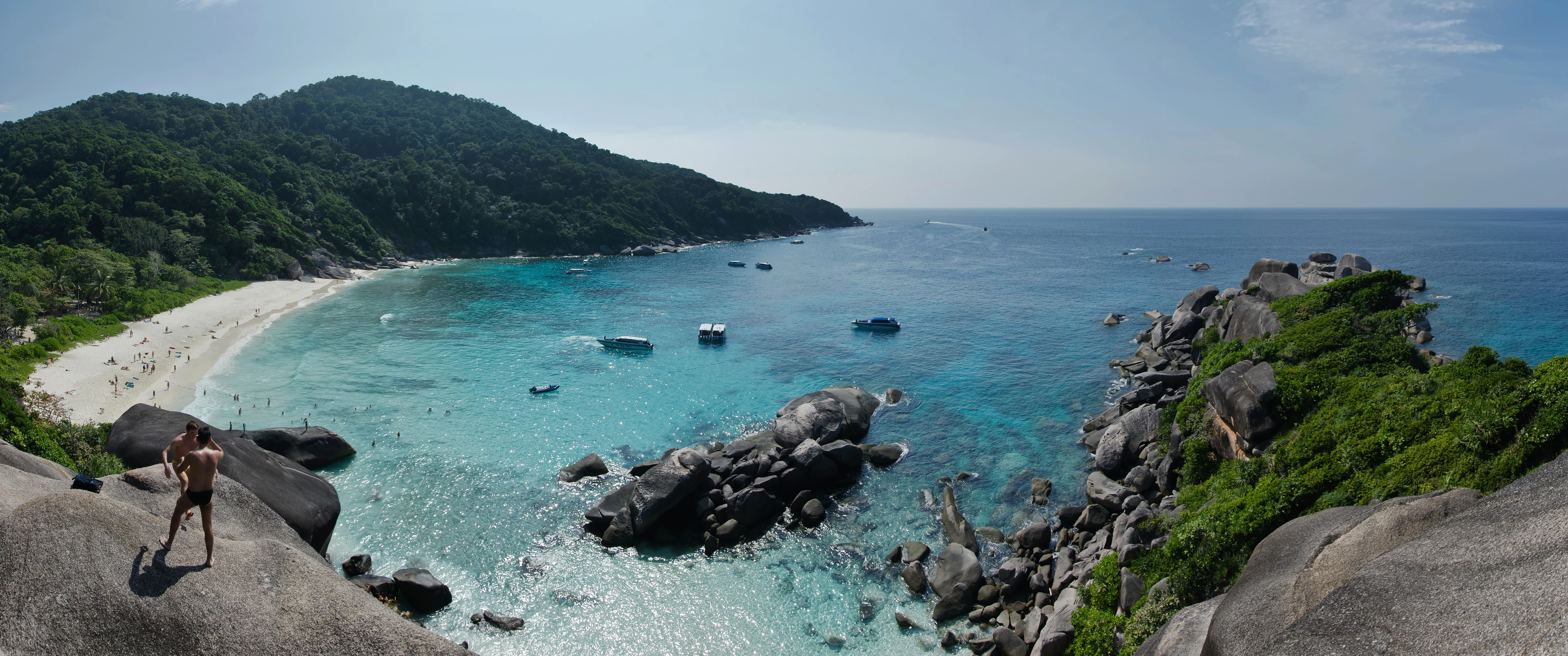 Panoramic landscape of Ko Similan beach with turquoise water, rocky outcrops, lush green hills, and a person standing on a rock