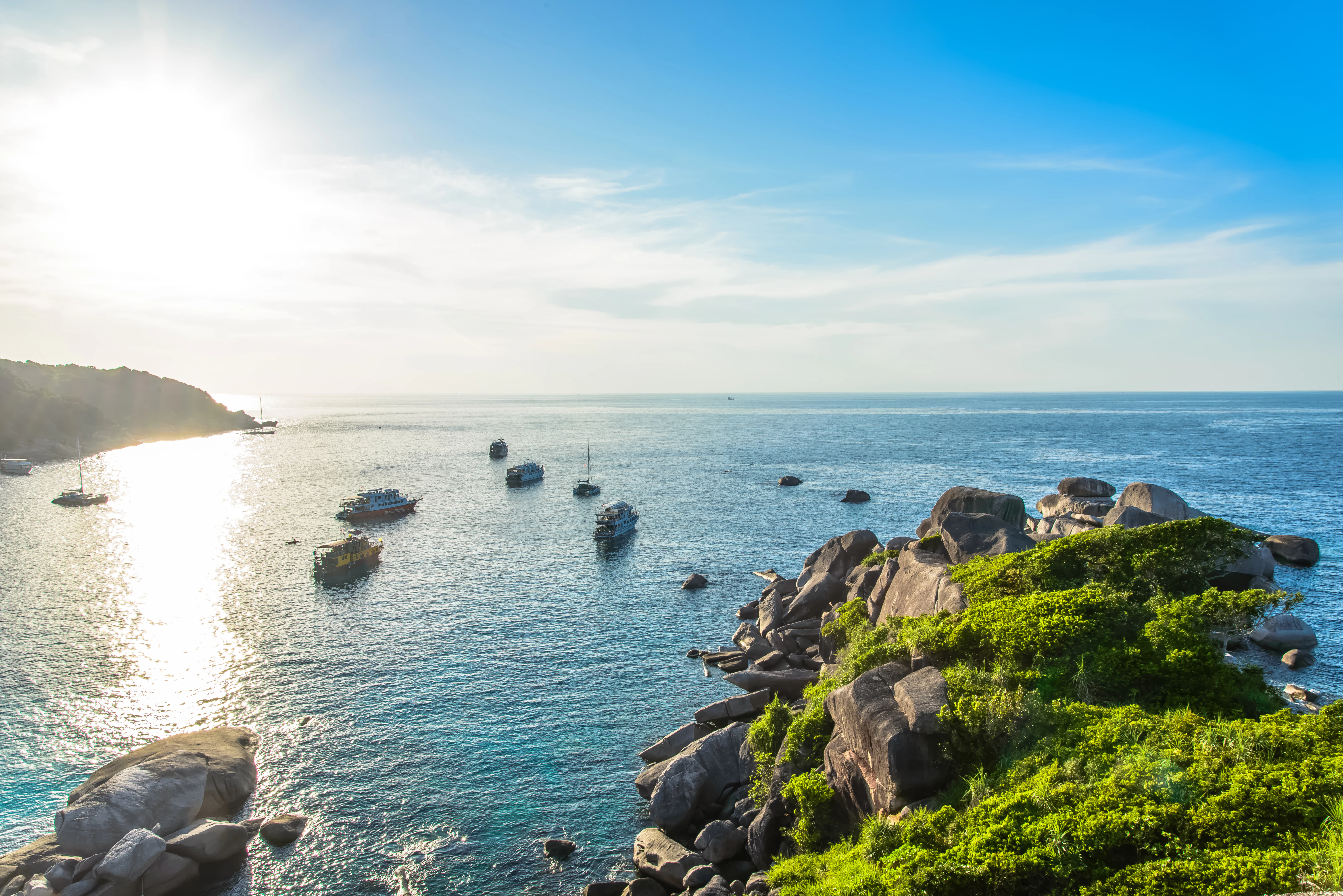 Rocky coastline with lush green vegetation overlooking the Andaman Sea with several boats anchored in the water under a clear blue sky