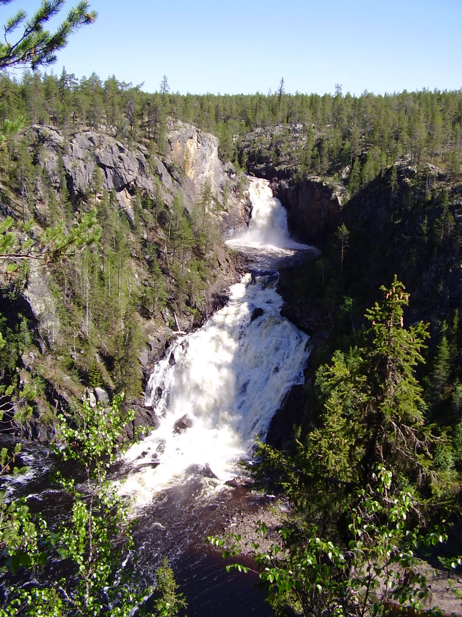 A waterfall cascading over rocks with dense forest in the background under a clear blue sky
