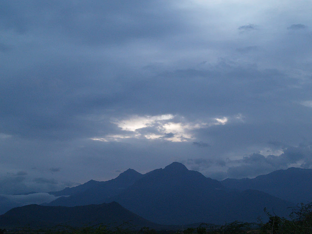 Dark blue sky with scattered clouds, sunlight breaking through, silhouetted mountain peaks, and foreground vegetation