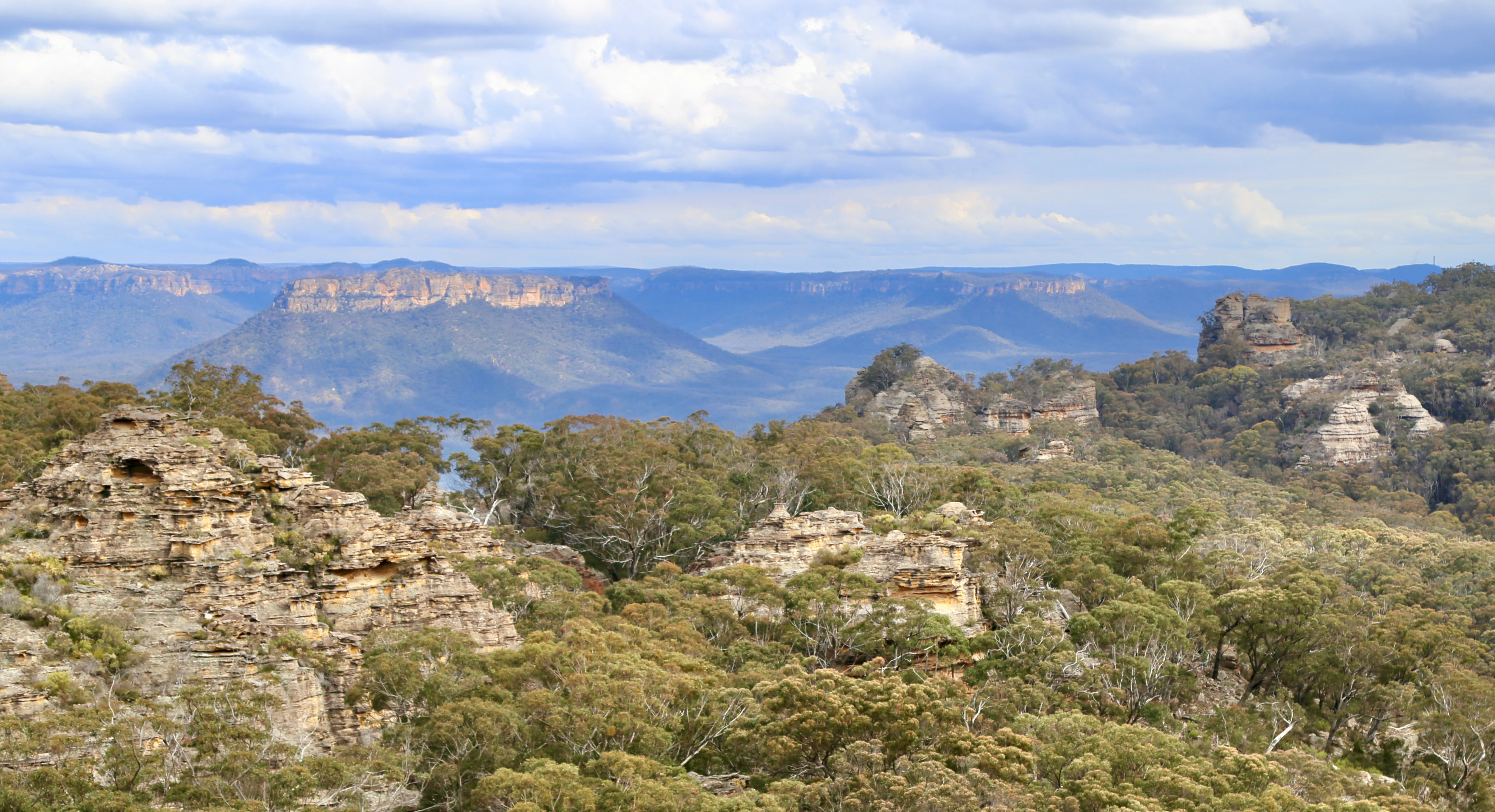 Panoramic landscape view of rugged sandstone pagodas in the foreground, with layers of blue mountain ranges and the flat-topped Patoneys Crown under a cloudy sky.