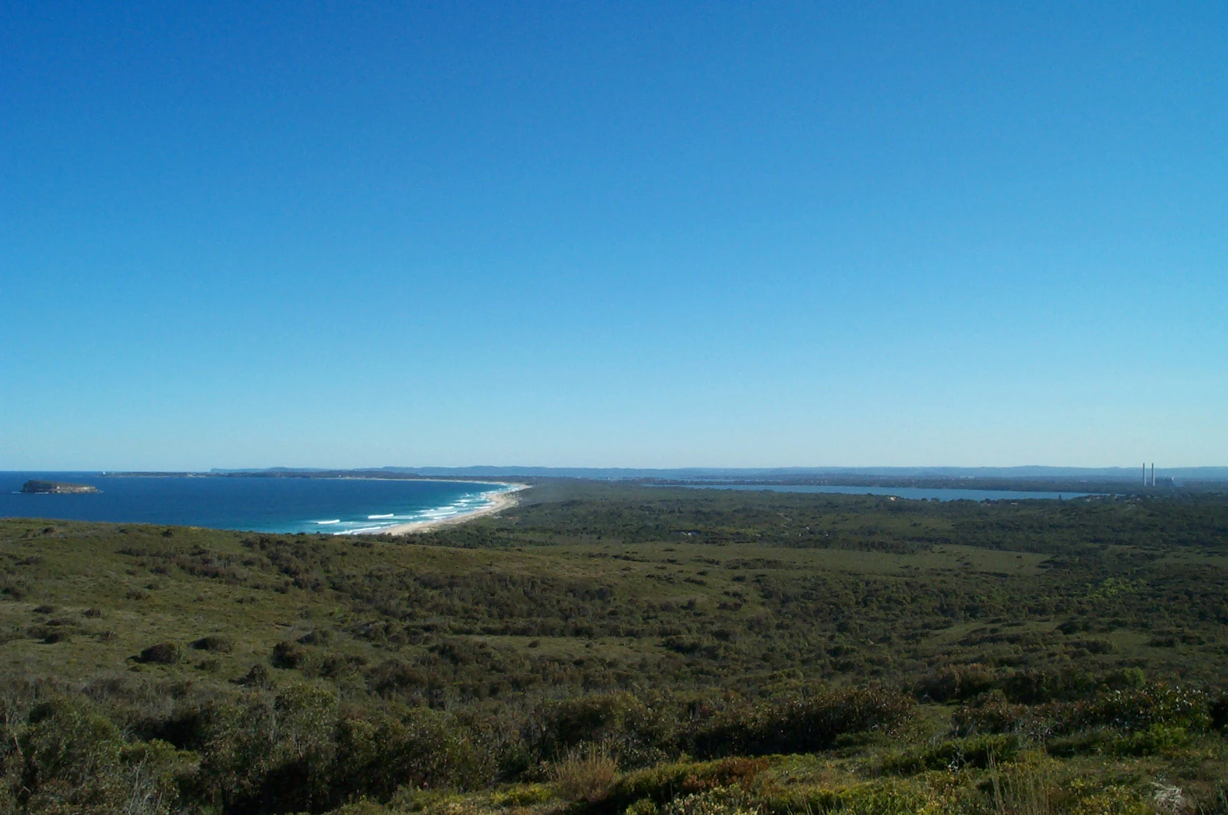 Panoramic landscape view featuring a coastal park with low scrubby vegetation in the foreground, stretching toward a beach along the blue ocean on the left, with a large lake visible inland.