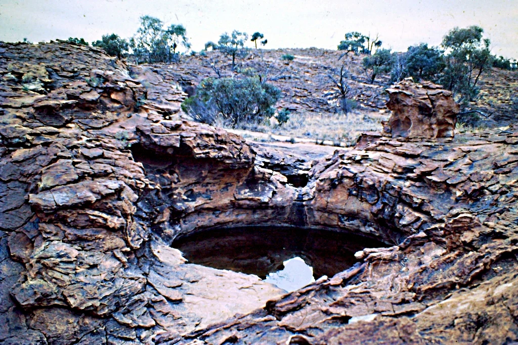 Circular waterhole surrounded by rugged layered rocks with sparse vegetation and distant trees