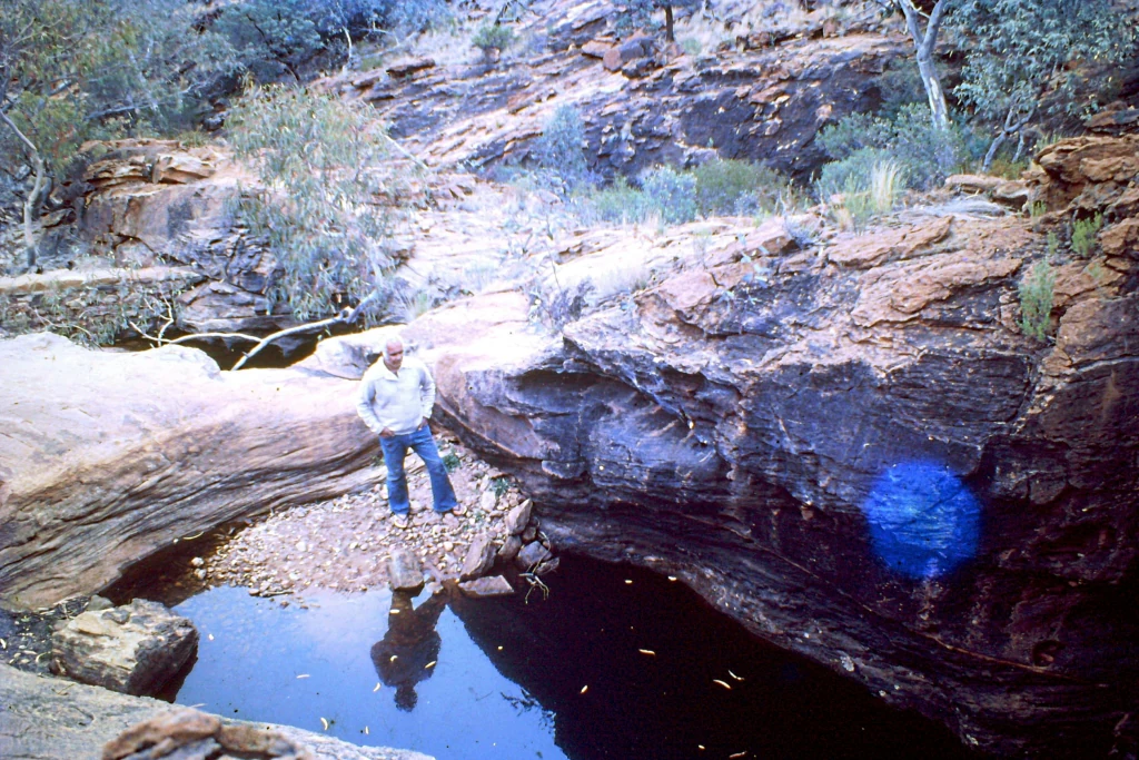 Person standing on rocky ledge by a waterhole surrounded by rugged red rock cliffs and sparse vegetation.