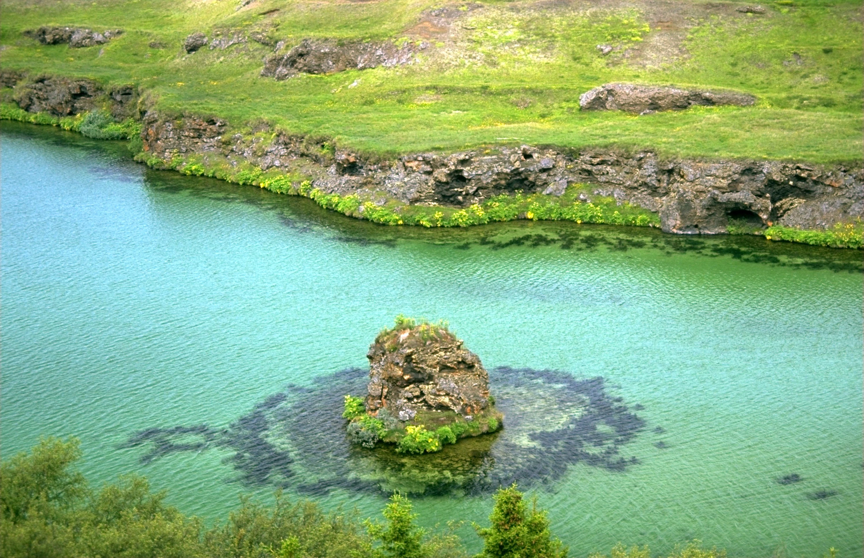 An elevated view of a small, rocky volcanic island surrounded by clear turquoise water, with a grassy, rocky shoreline in the background.