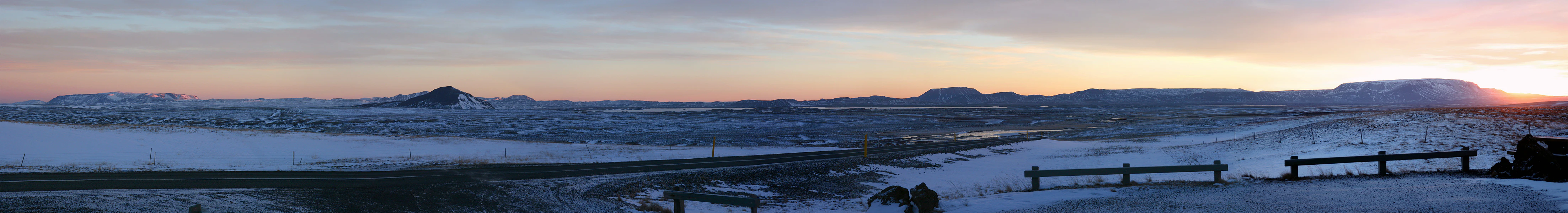 A wide panoramic view of a snow covered volcanic landscape in Iceland under a soft sunrise sky, with several distinct mountains and a wooden guardrail in the foreground.