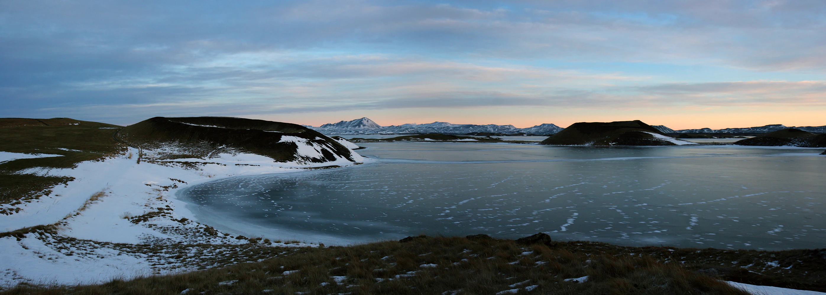 A panoramic view of Mývatn lake in winter, with snow-covered volcanic pseudocraters on the shoreline and distant snow-capped mountains under a soft twilight sky.