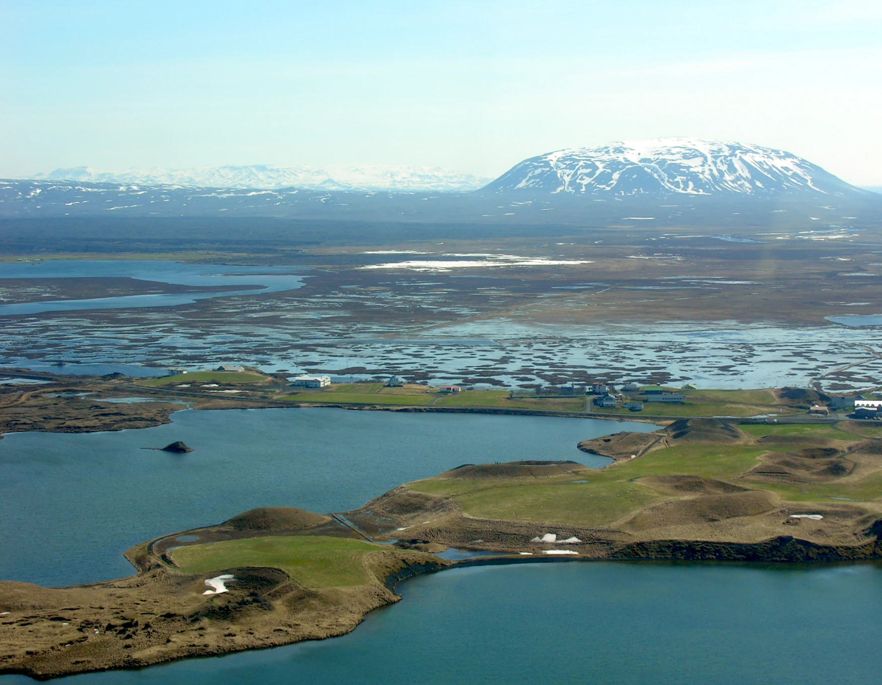 An aerial perspective of Mývatn showing a large lake with small, grassy volcanic pseudo craters on the shore and a snow-capped mountain in the distant background.