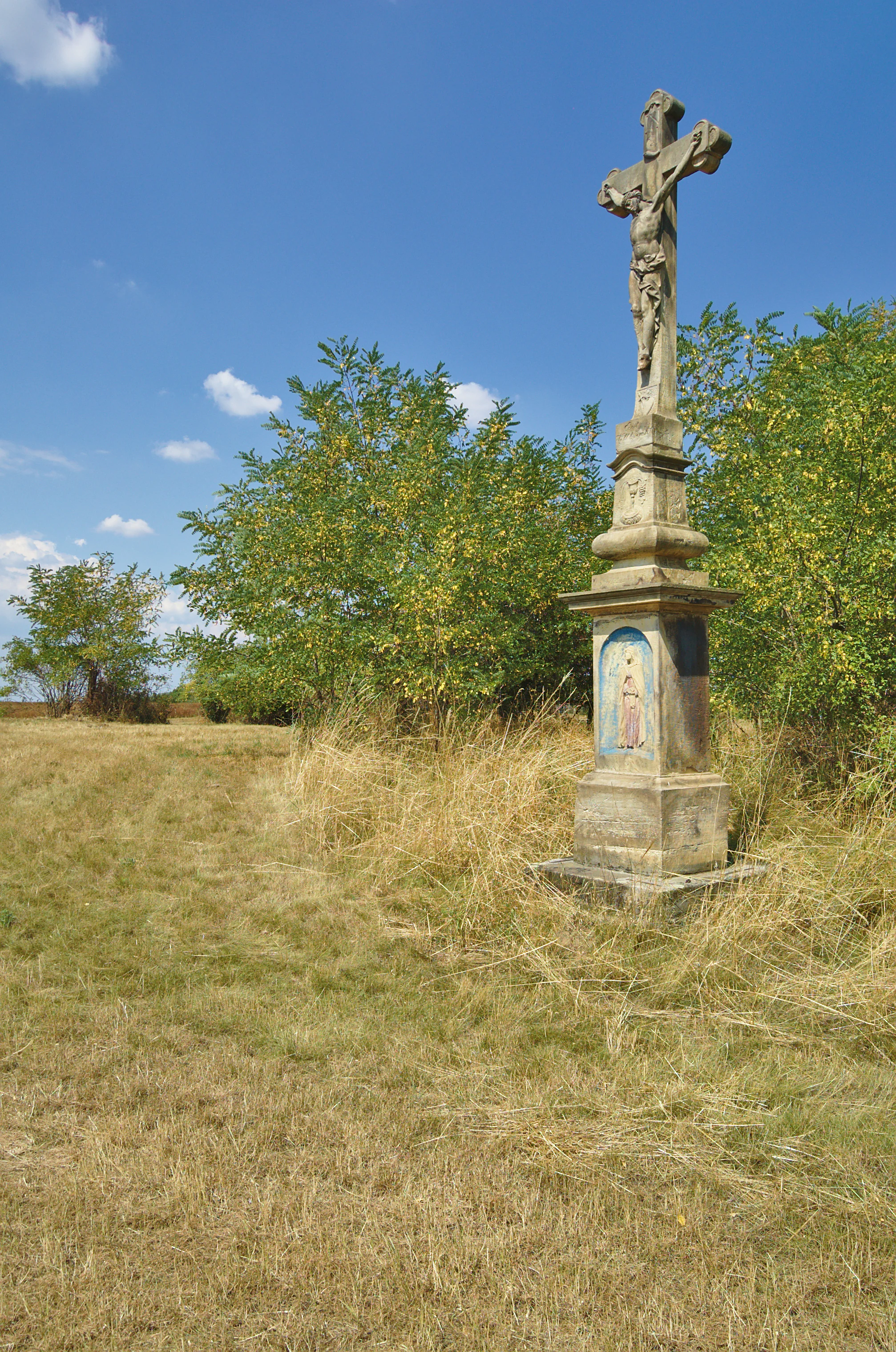 An old stone crucifix featuring a sculpture of Jesus Christ, standing in a dry, grassy field surrounded by shrubs under a blue sky.