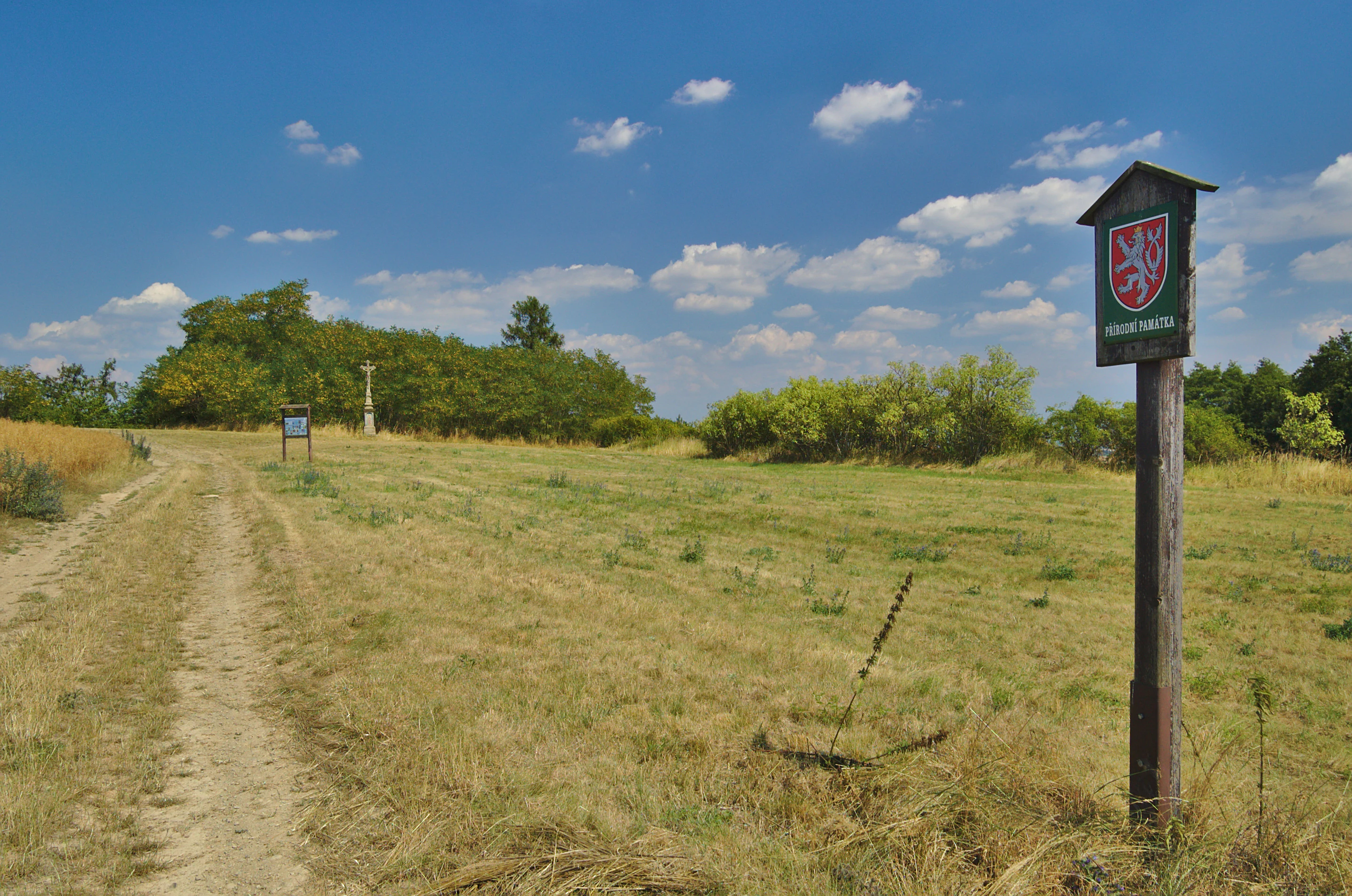 A wooden sign featuring the Czech coat of arms stands in a grassy field at the Na hůrkách natural monument, with a dirt path leading towards trees in the distance.