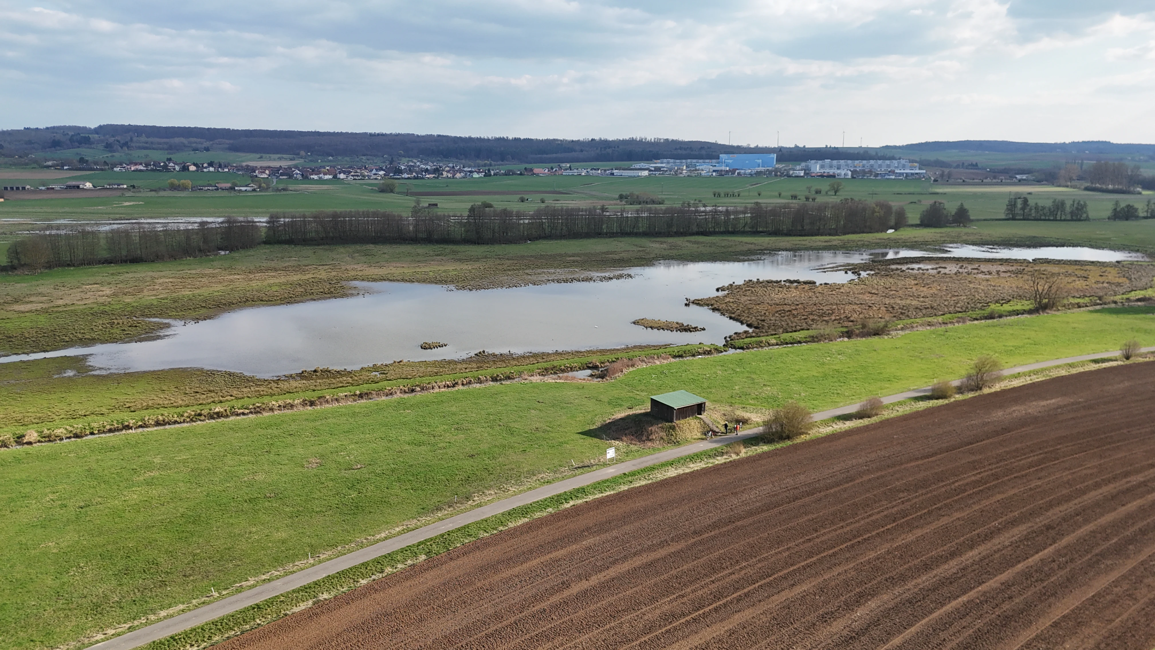 An aerial landscape view shows a nature reserve with a wetland pond surrounded by green fields, a small structure, and tilled farmland in the foreground.