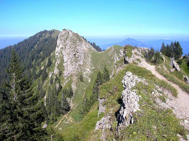 A jagged rocky mountain peak rises sharply above a grassy, forested ridge with a hiking trail running along the spine.