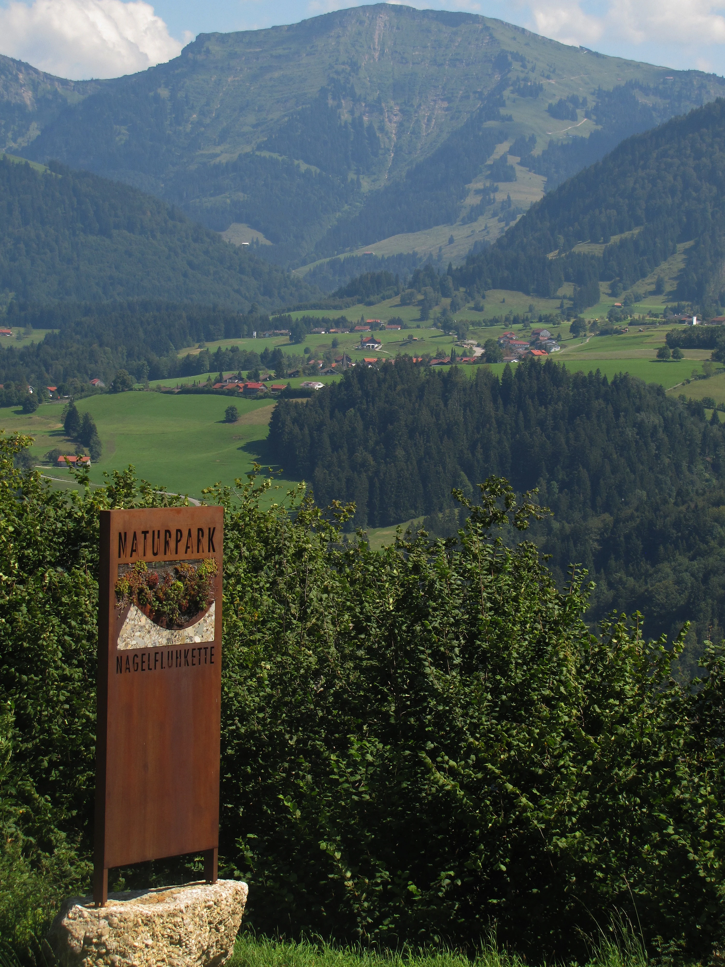 A tall, brown metal park entrance sign stands in the foreground, displaying the text Naturpark Nagelfluhkette; in the background, a large mountain rises above green meadows and forested slopes.