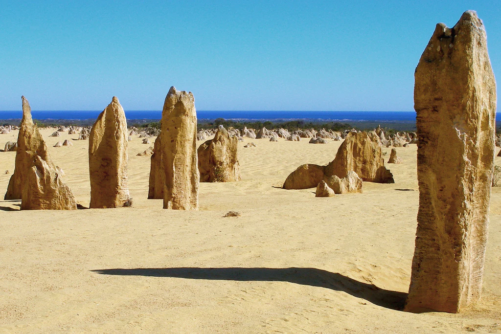 Limestone pillars in a sandy desert landscape with the Indian Ocean on the horizon under a clear blue sky