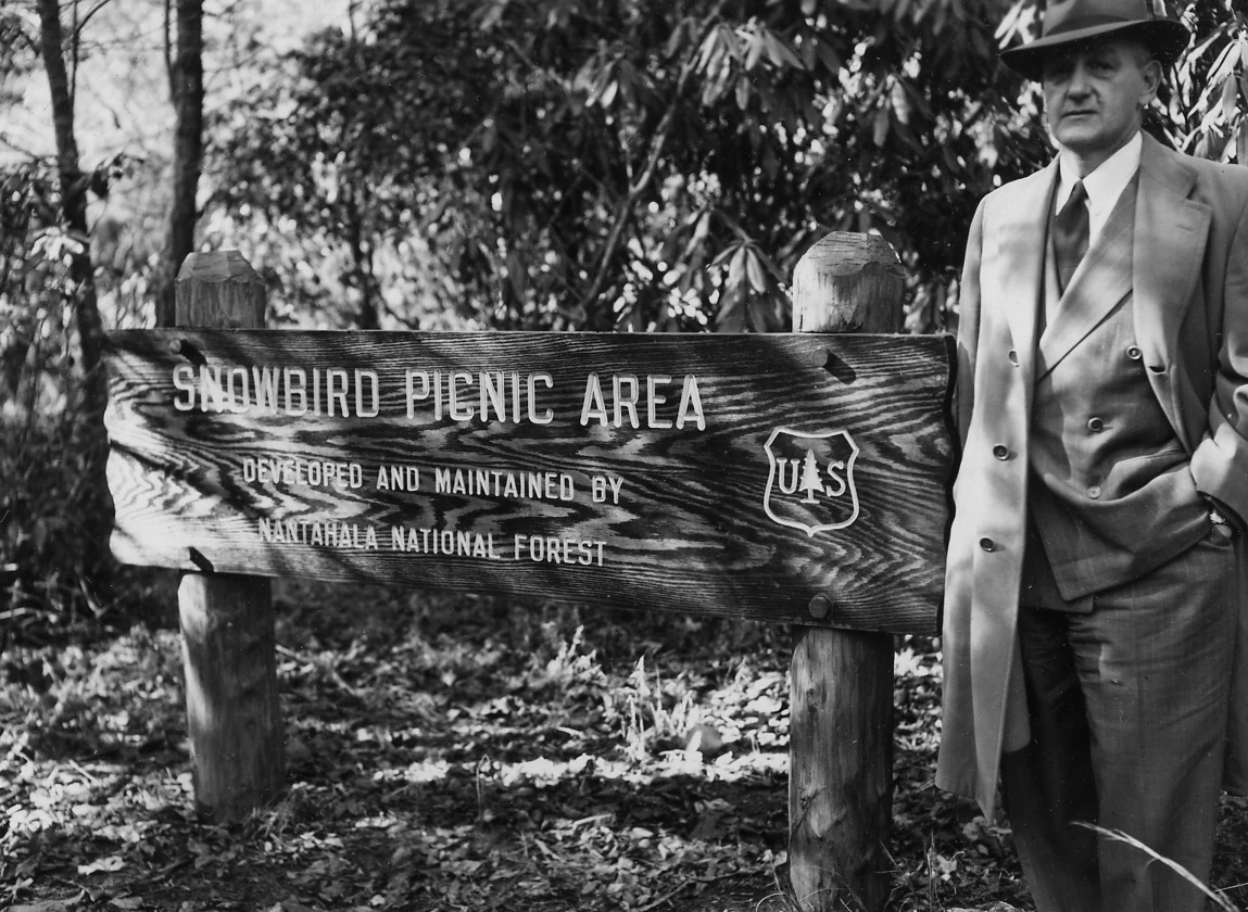 A black and white photograph showing a man in a suit and fedora standing next to a weathered wooden sign labeled Snowbird Picnic Area, Developed and maintained by Nantahala National Forest, featuring a US Forest Service shield logo.