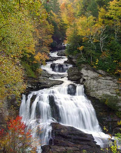 A multi-tiered waterfall cascading down dark rocky ledges, surrounded by dense trees with autumn foliage in shades of gold, orange, and red.