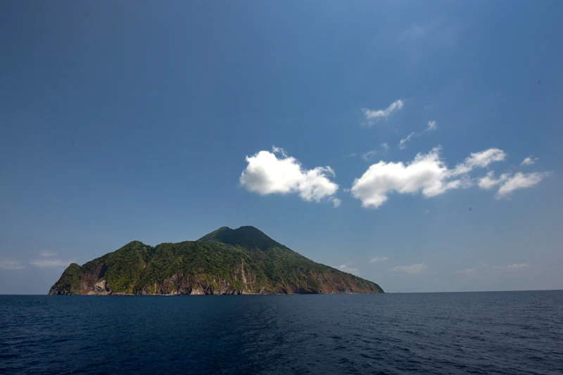 A wide shot of a lush, green volcanic island rising steeply from the deep blue ocean under a clear sky with scattered white clouds.