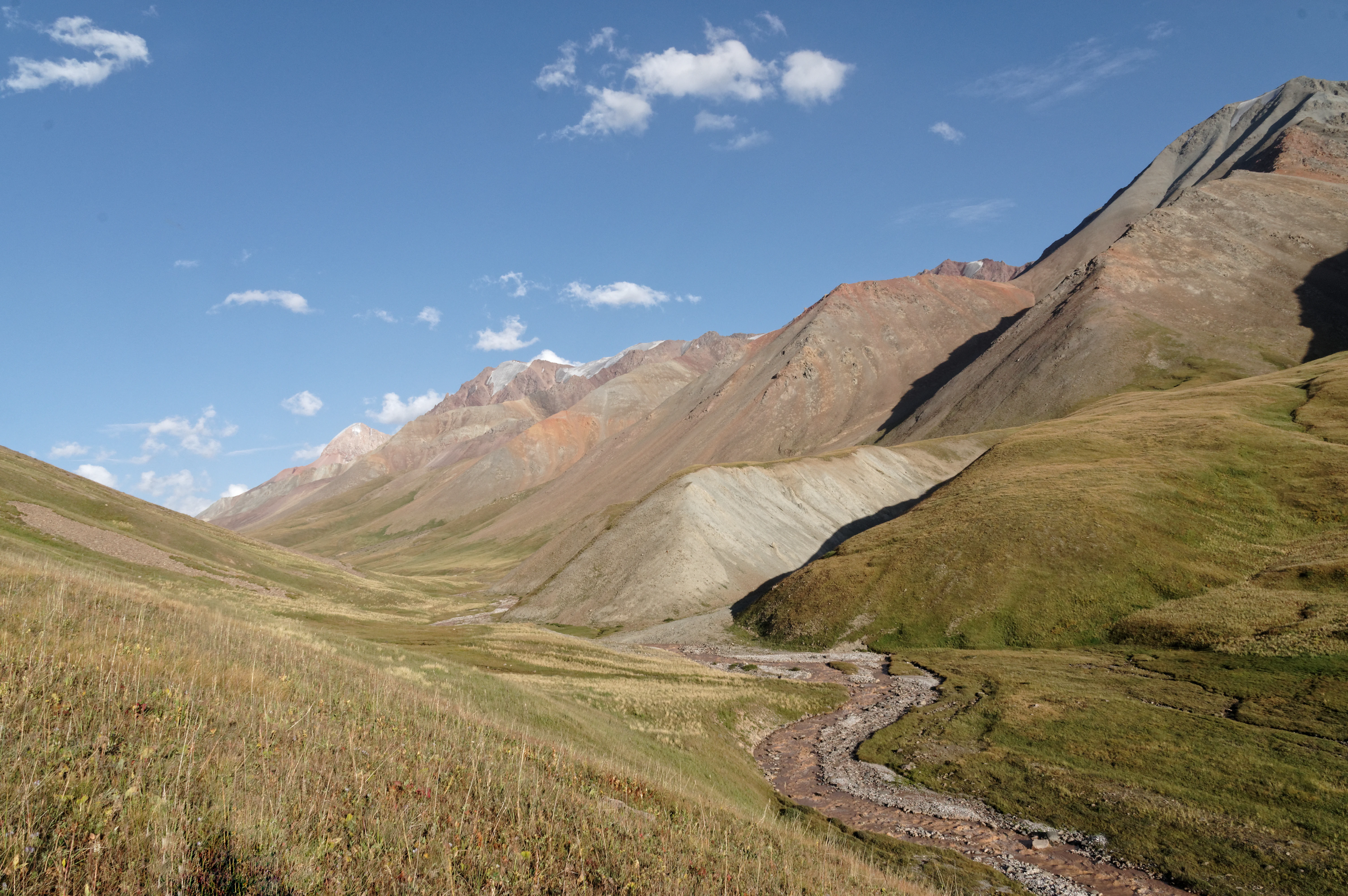 A wide landscape view of a mountain valley in Naryn Nature Reserve featuring dry grassy slopes, rocky peaks, and a narrow river winding through the center under a clear blue sky.