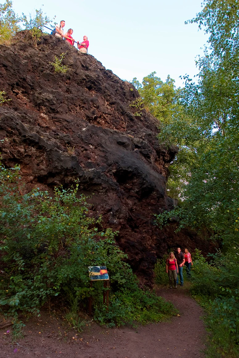 A group of people standing on top of a dark, jagged volcanic rock cliff with others walking on a trail below past an information sign.