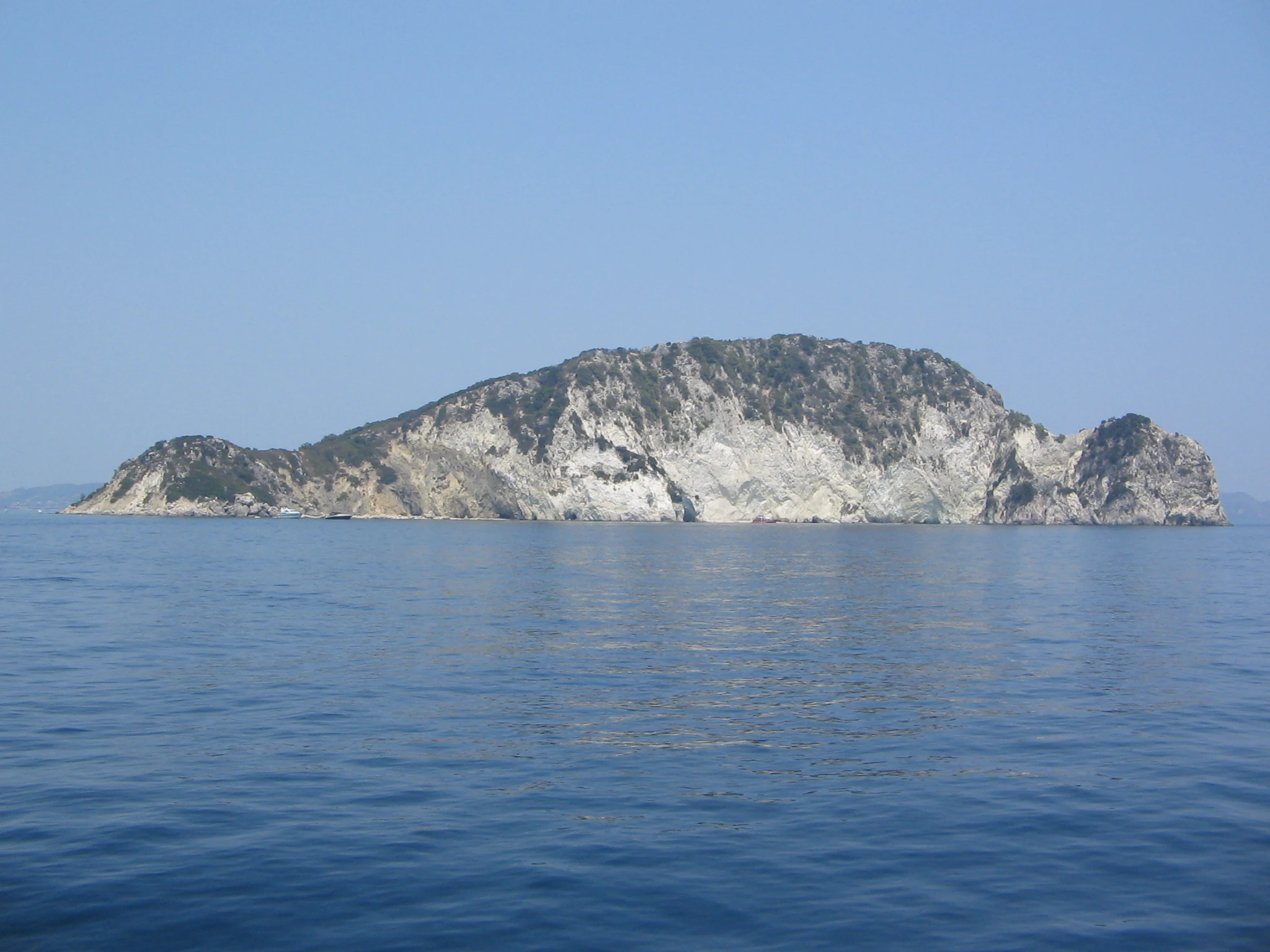 An island with rocky terrain and sparse vegetation visible in the Ionian Sea under clear blue sky