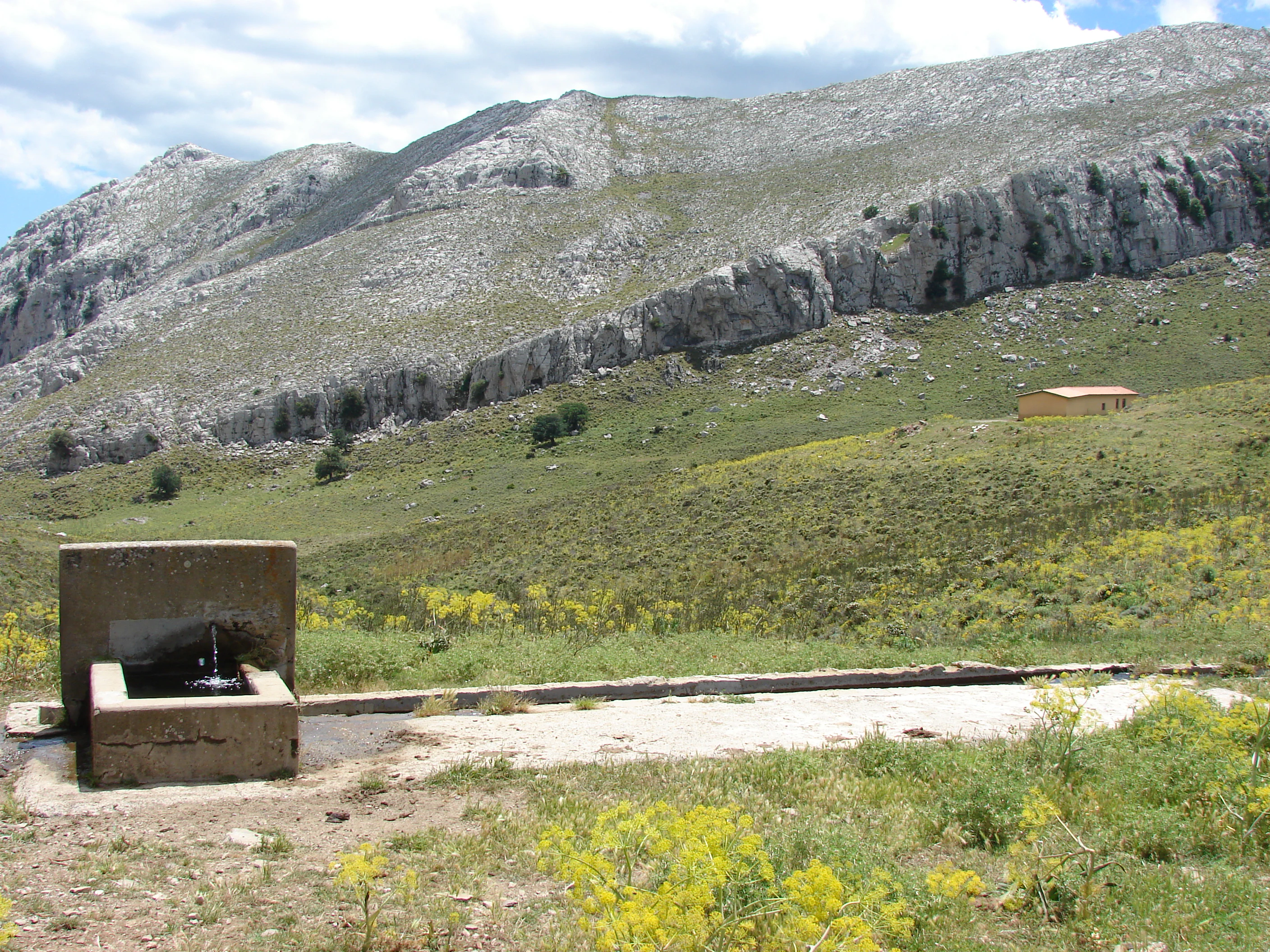 Concrete water fountain structure on a grassy hillside with rocky mountains in the background and a small building