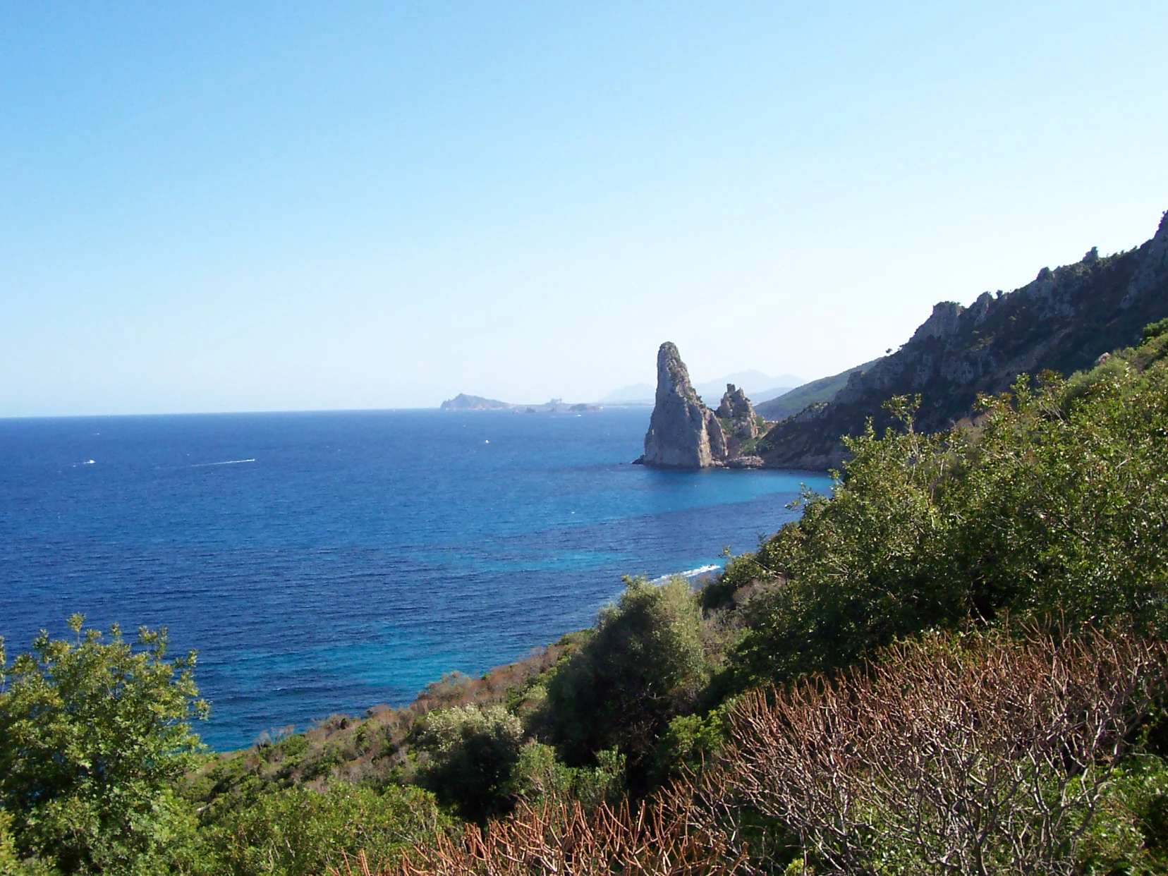 Coastal landscape with blue sea, Pedra Longa rock formation, green hills, and clear sky