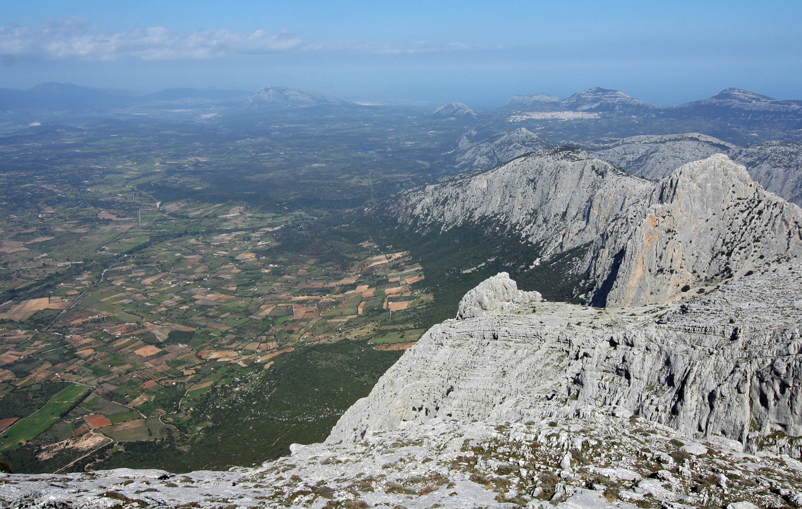 Rocky mountain peak overlooking valleys, forests, and distant coastline under a clear sky