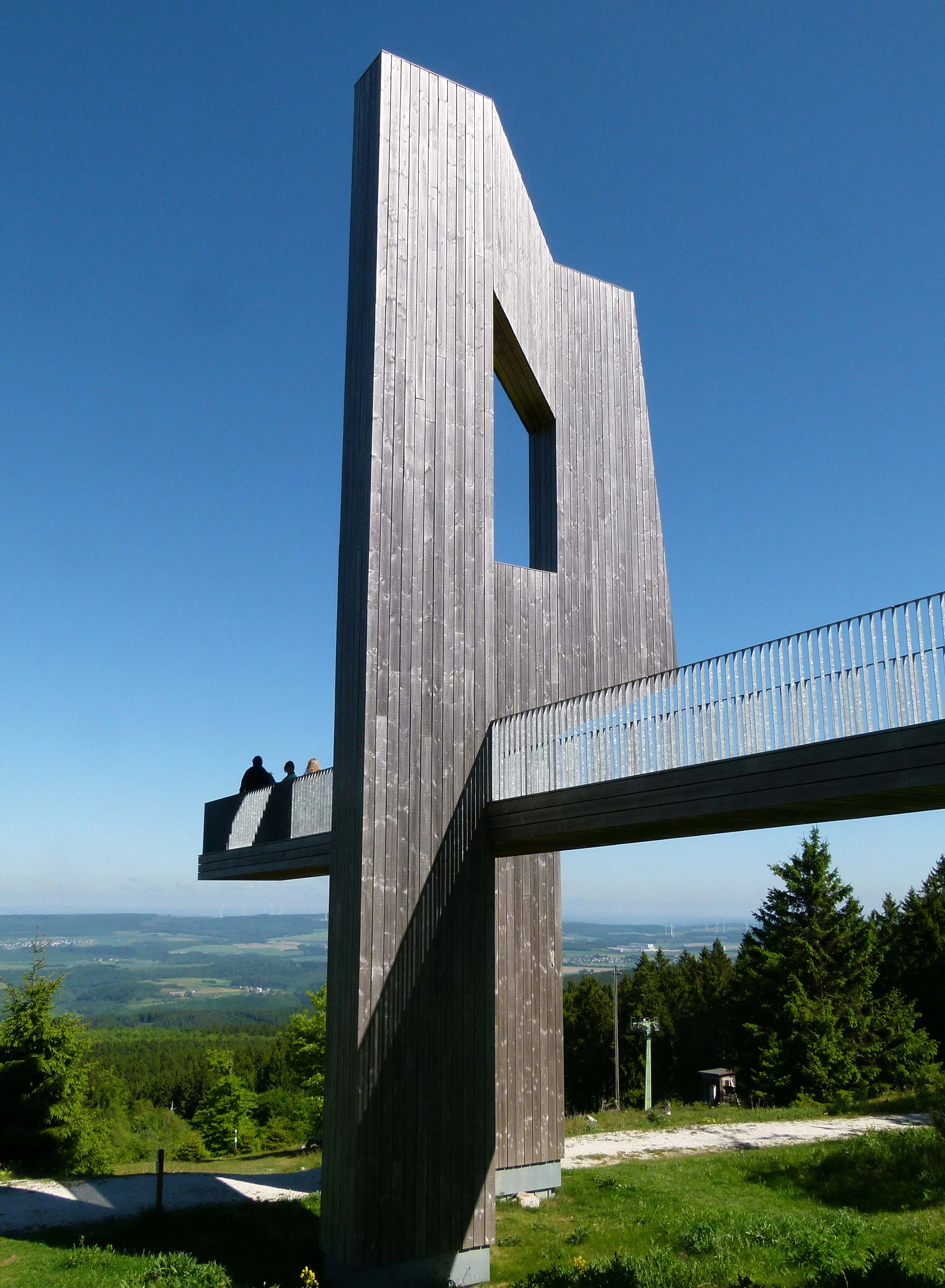A tall, vertical wooden sculpture with a cut-out window, integrated with a metal-railed observation platform at the Erbeskopf mountain peak, overlooking a green forest valley under a clear blue sky.