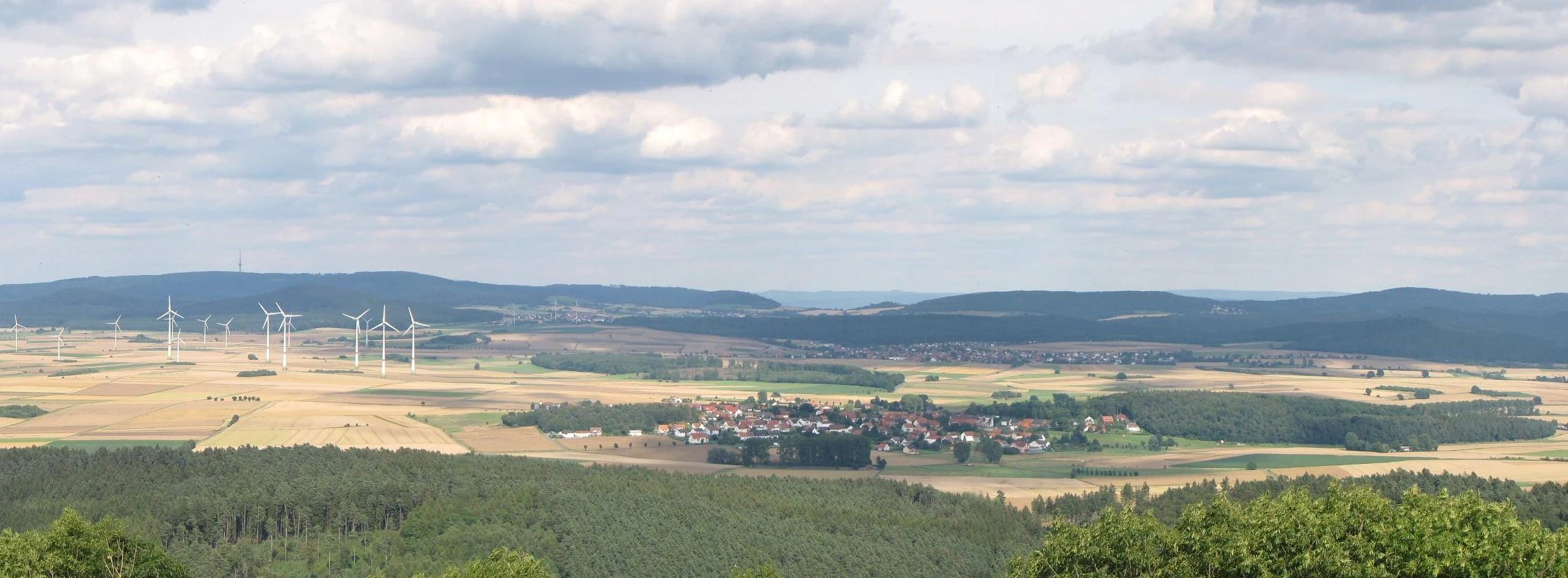 A wide, elevated landscape view featuring rolling hills covered in dense forest, interspersed with agricultural fields and a small village, with wind turbines visible in the middle distance under a cloudy sky.