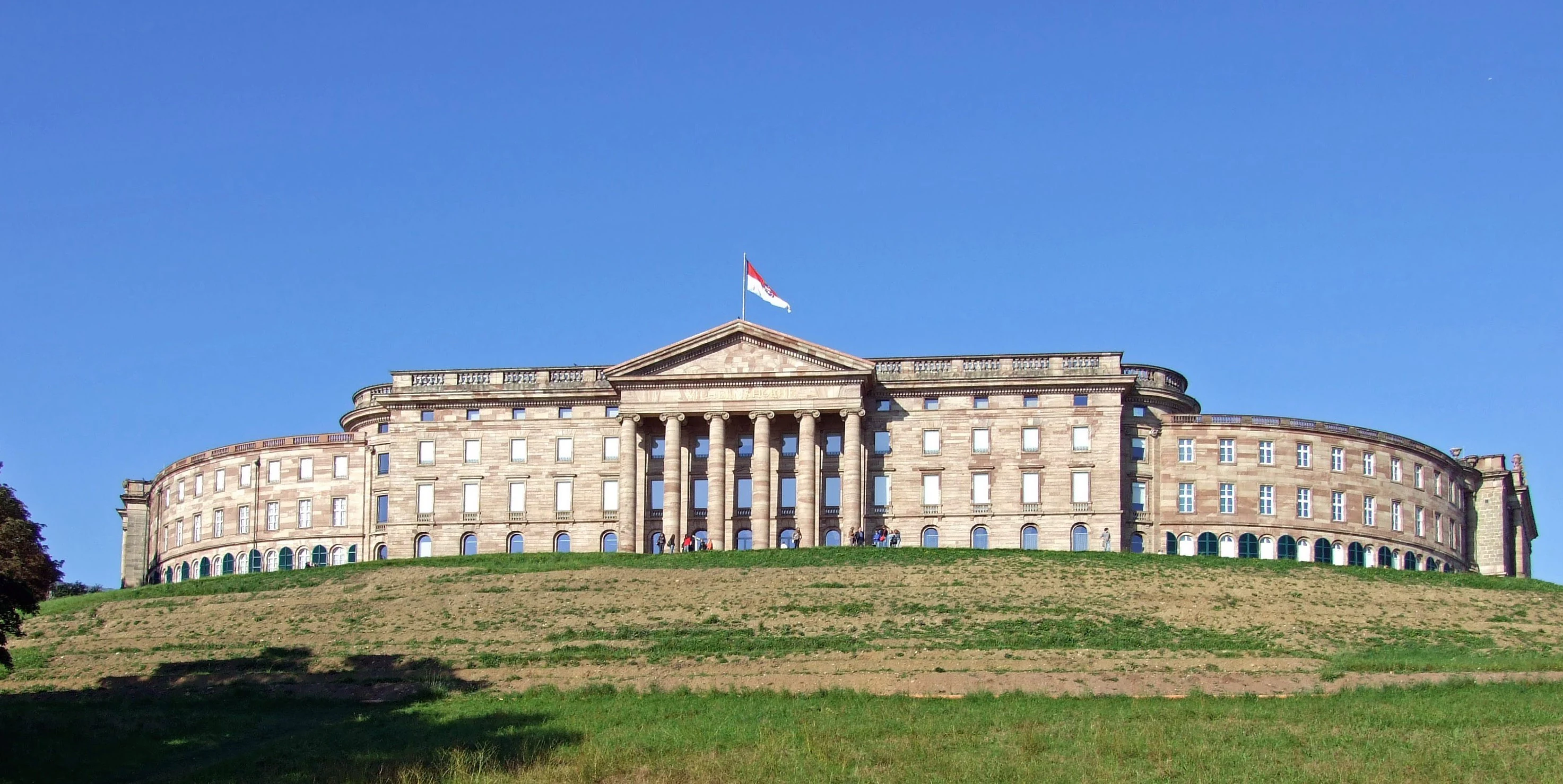 A wide, eye-level shot of the large, neoclassical Schloss Wilhelmshöhe palace, showing its curved wings and central pedimented portico atop a grassy hill under a clear blue sky.