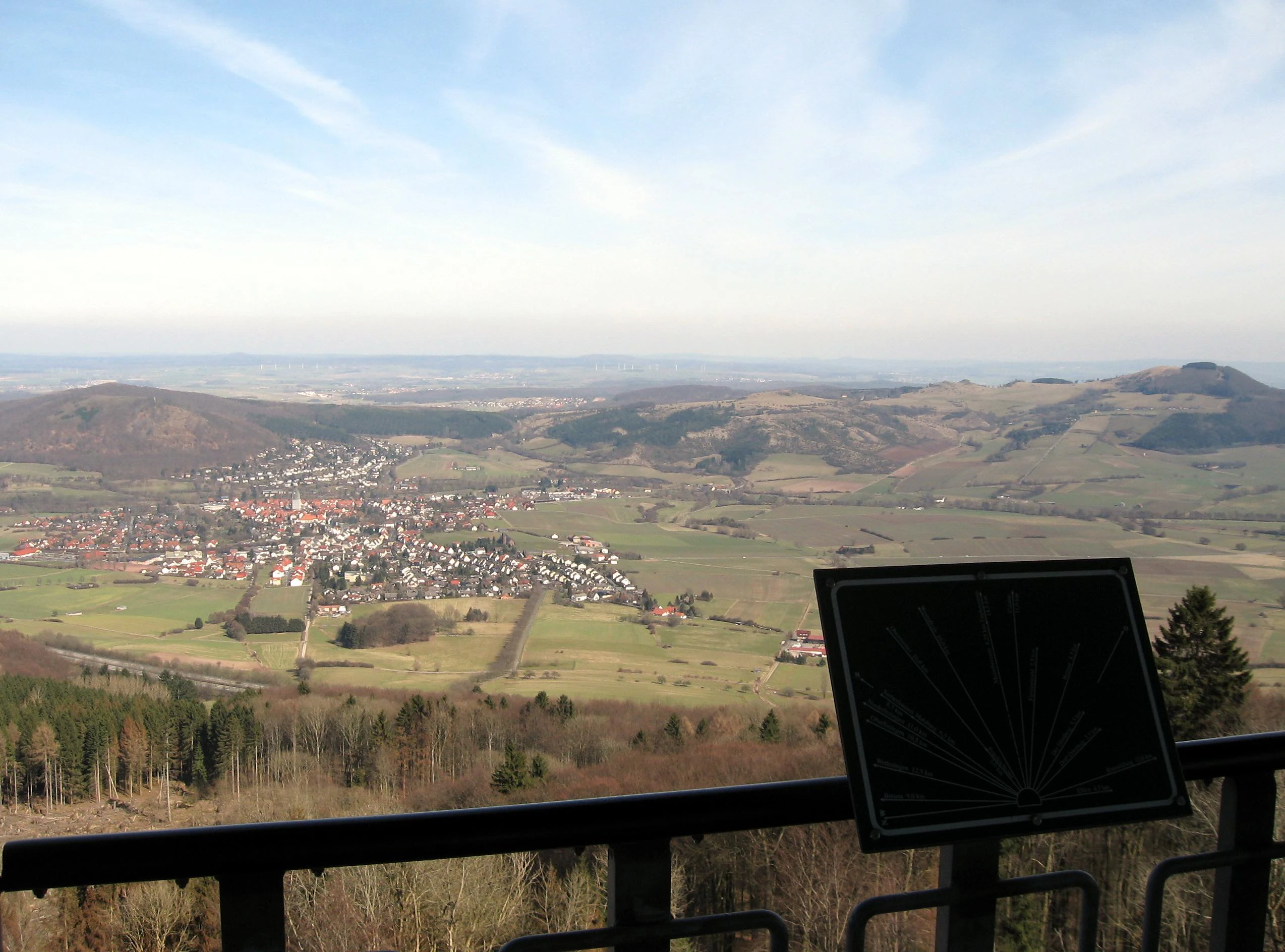 High angle view from an observation deck looking out over a small town nestled in a valley, with rolling forested hills in the background and a black metal orientation map on a railing in the foreground.