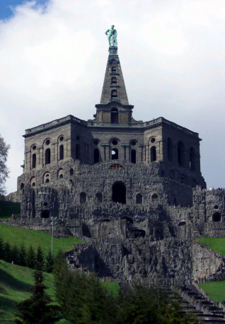 A low-angle, full view of the stone Hercules Monument, featuring an octagonal structure topped by a spire and a statue, built upon a rugged grotto-like foundation surrounded by green parkland.