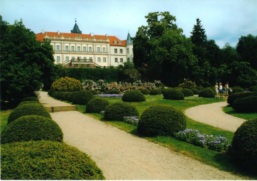 A wide view of Schloss Wiesenburg, a large historic building with a red roof, viewed from behind manicured hedges and winding gravel paths in a garden.