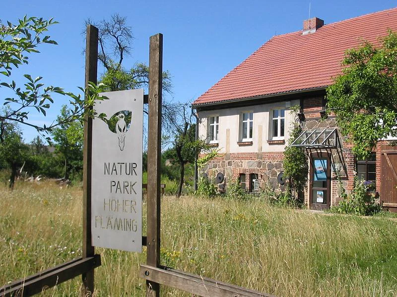 A sign on wooden posts in the foreground identifies the Naturpark Hoher Fläming, with a traditional brick and stone building visible in the background.
