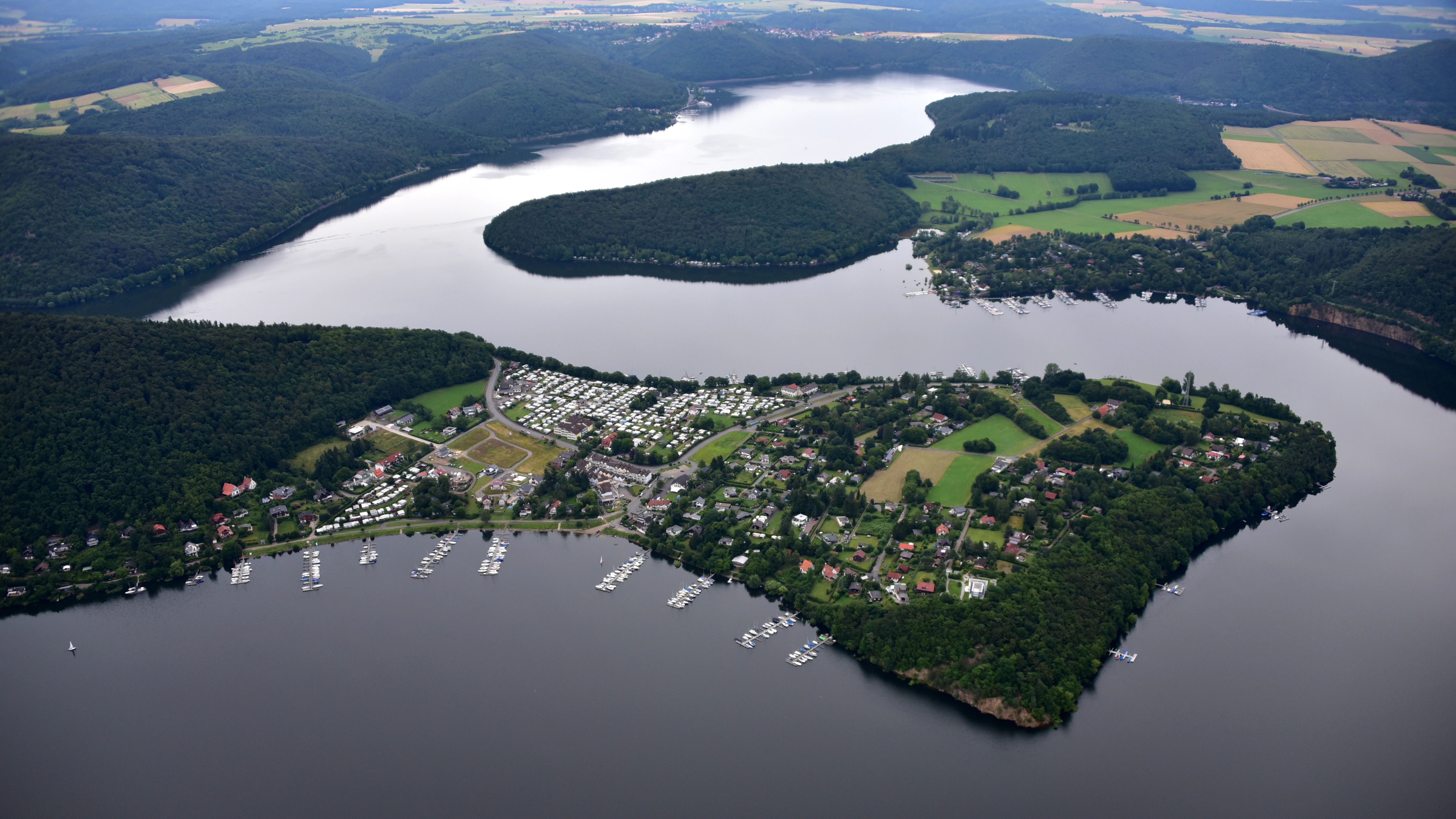 An aerial view of a lake and a large peninsula with a residential area, docks, and boats, surrounded by dense green forest.