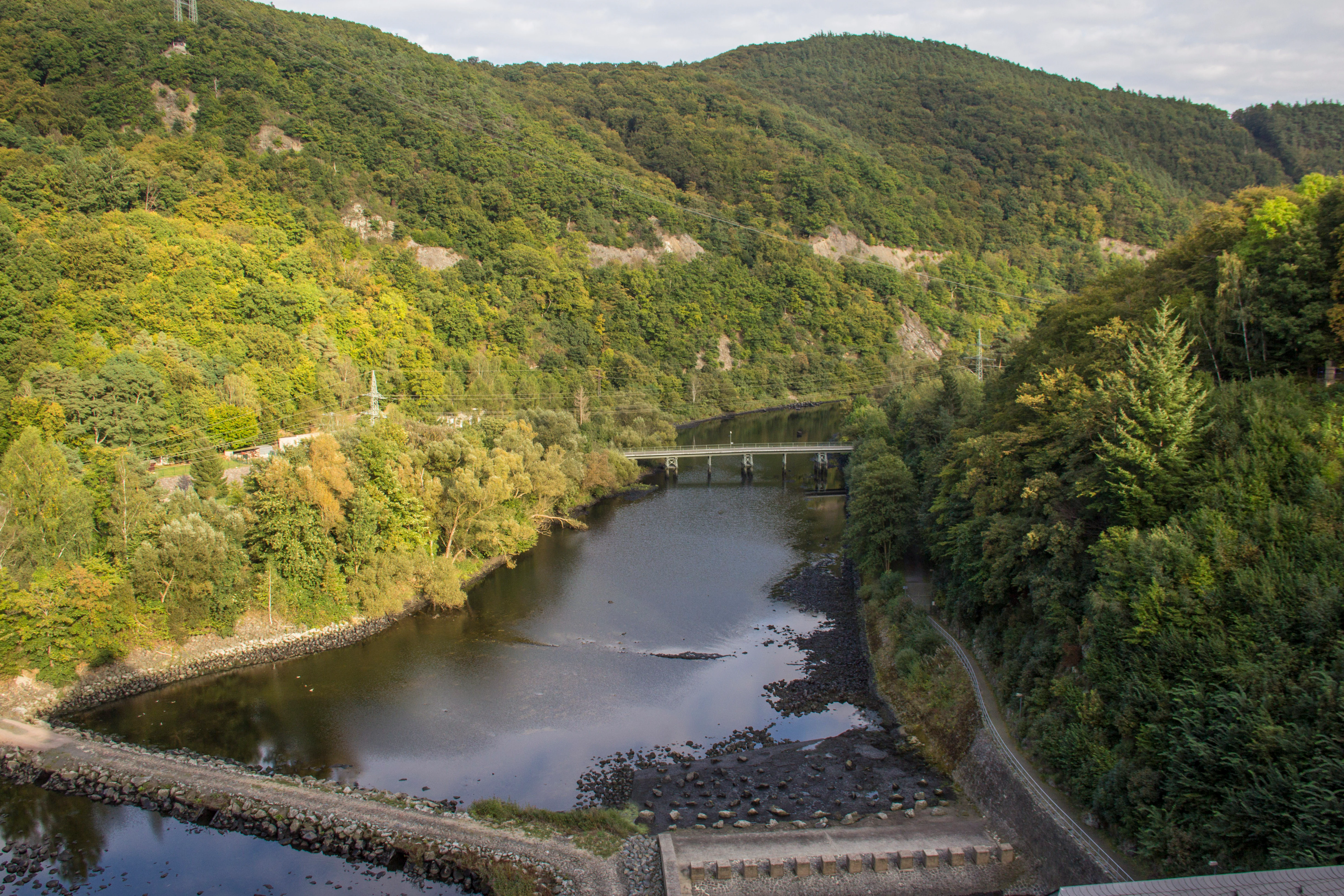 An elevated view looking down into a forested river valley, featuring a calm river winding through dense green trees and a small concrete dam structure.