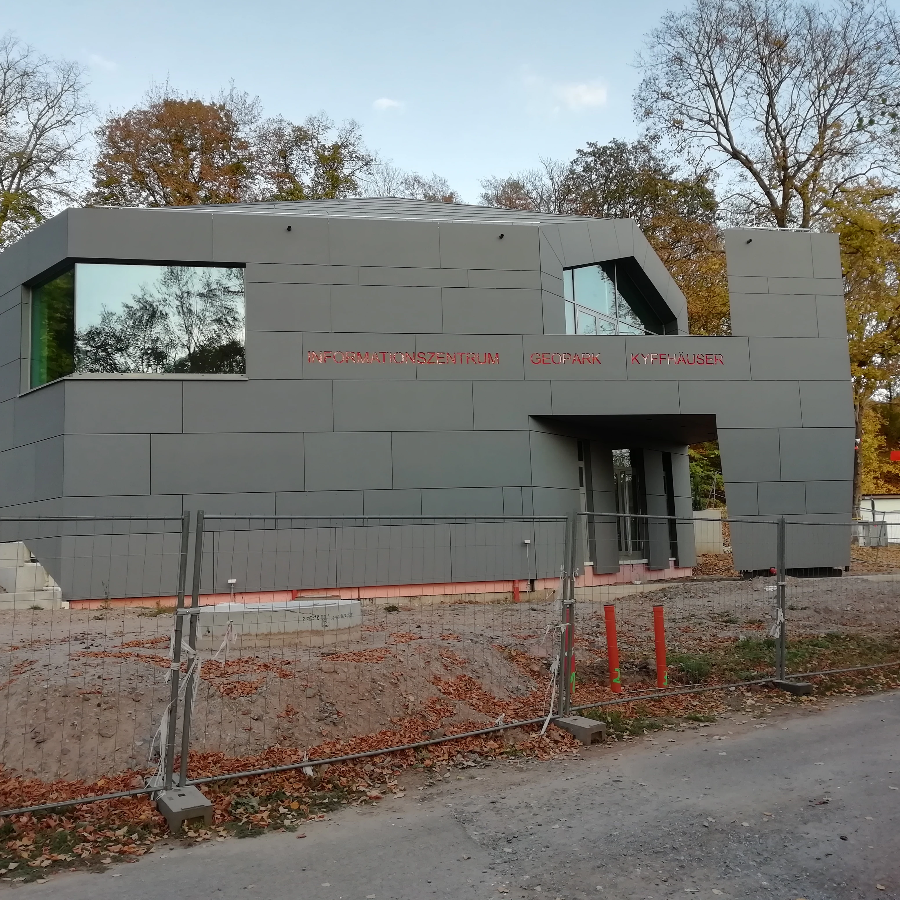 Un edificio moderno con paneles grises y grandes ventanales geométricos sirve como centro de información para visitantes de la Barbarossahöhle, visto desde el otro lado de una zona de grava con vallas de obra.