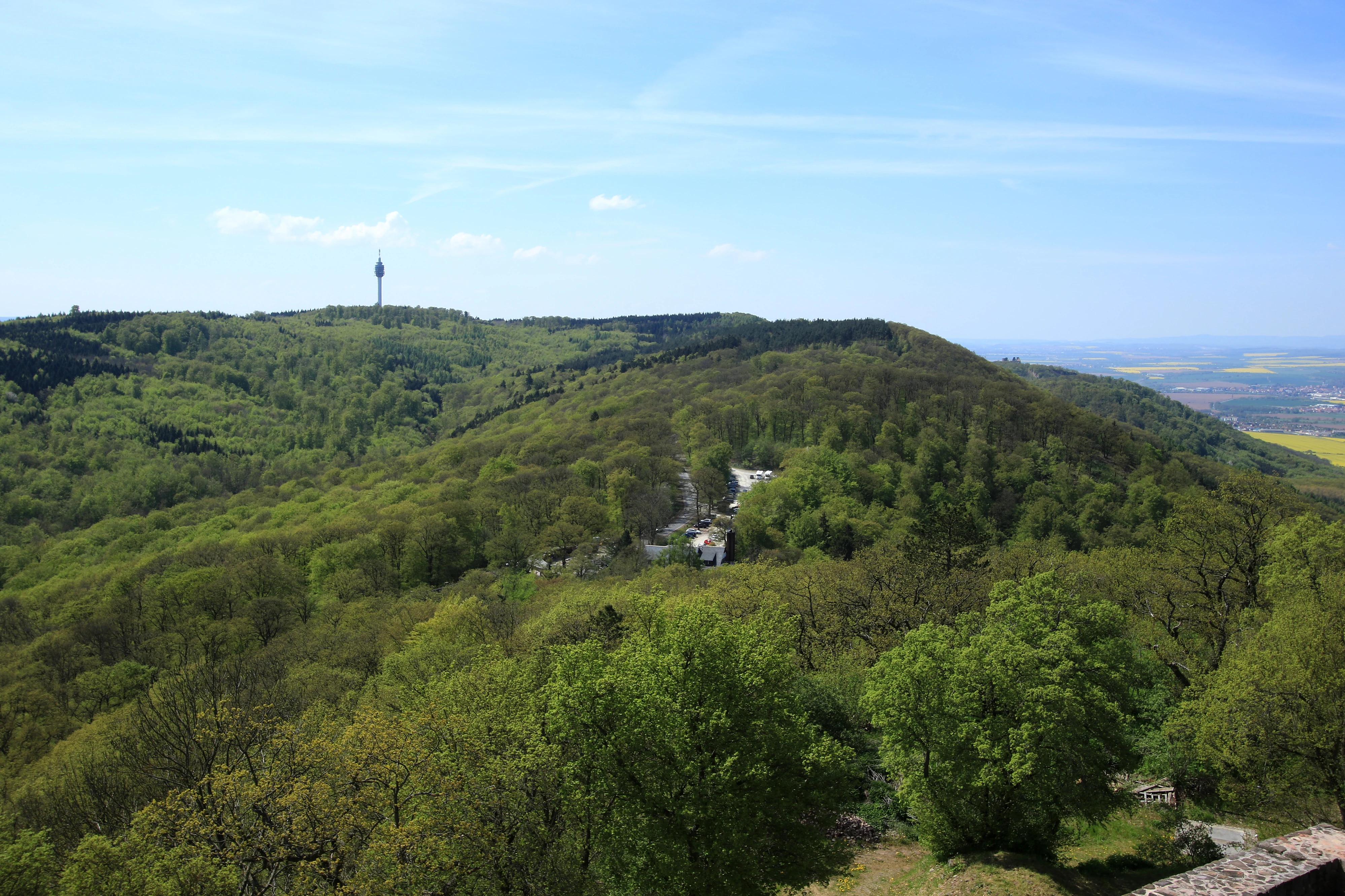 Plano general del paisaje con colinas verdes y boscosas bajo un cielo azul, con una torre de telecomunicaciones alta visible en una cresta lejana.
