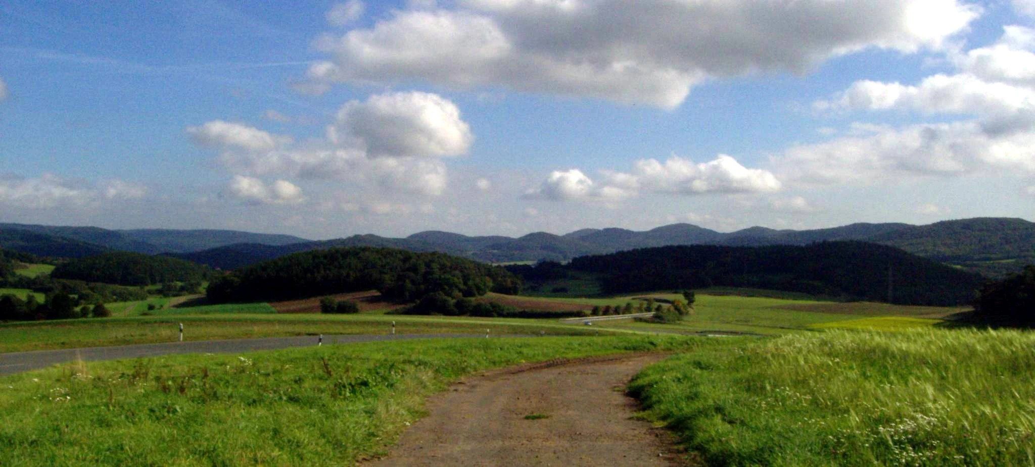 A wide, panoramic landscape view featuring rolling green hills, forested slopes, and a dirt path leading through meadows in the foreground under a blue sky with scattered white clouds.
