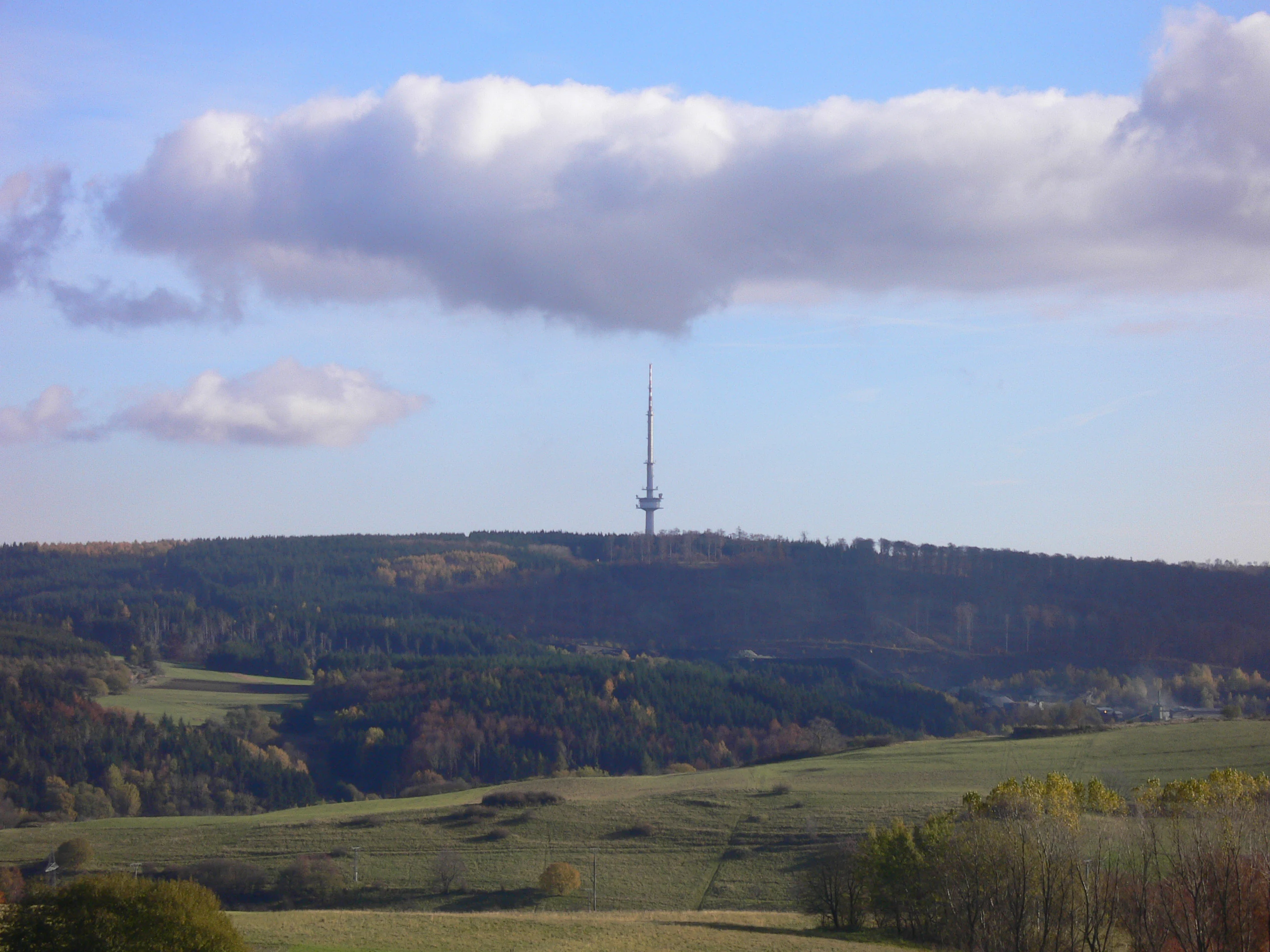 A distant view of a tall television transmitter tower standing on a forested hill, surrounded by rural pastures and fields under a sky with scattered clouds.