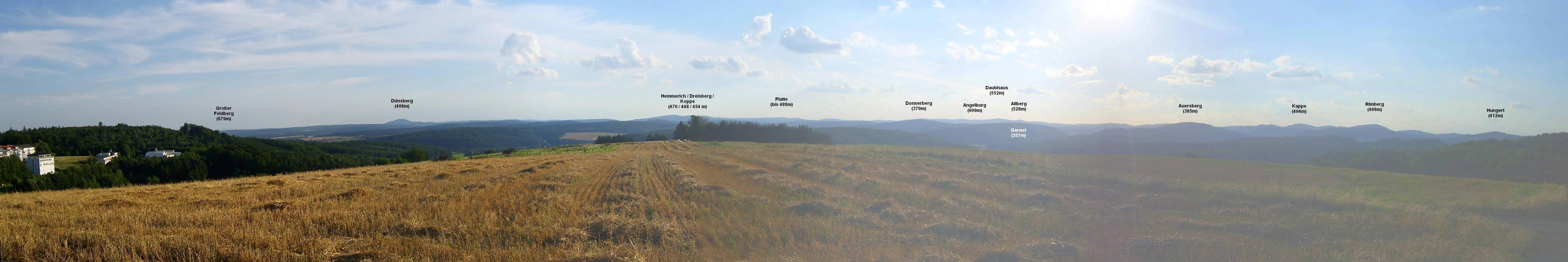 A wide panoramic photograph showing golden fields in the foreground transitioning into soft rolling hills and ridges under a blue sky, with small text labels overlaid on the distant landscape identifying various peaks.